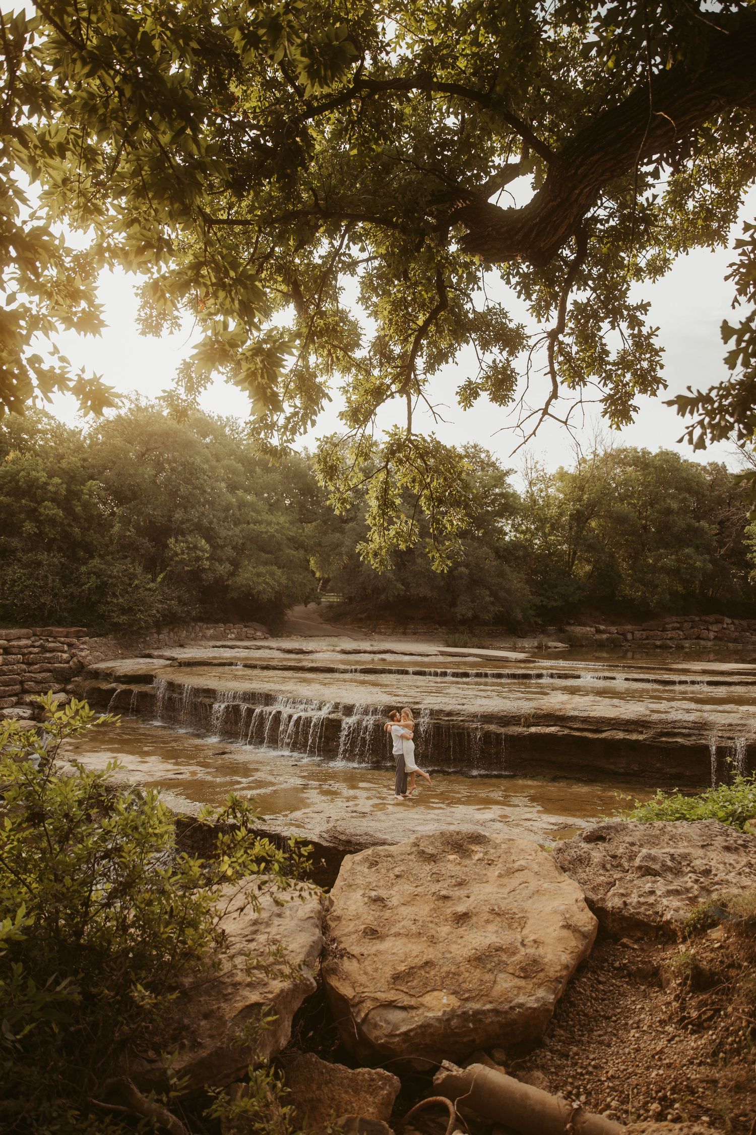 Couple embracing on rocky waterfall steps surrounded by lush green trees in golden hour light.
