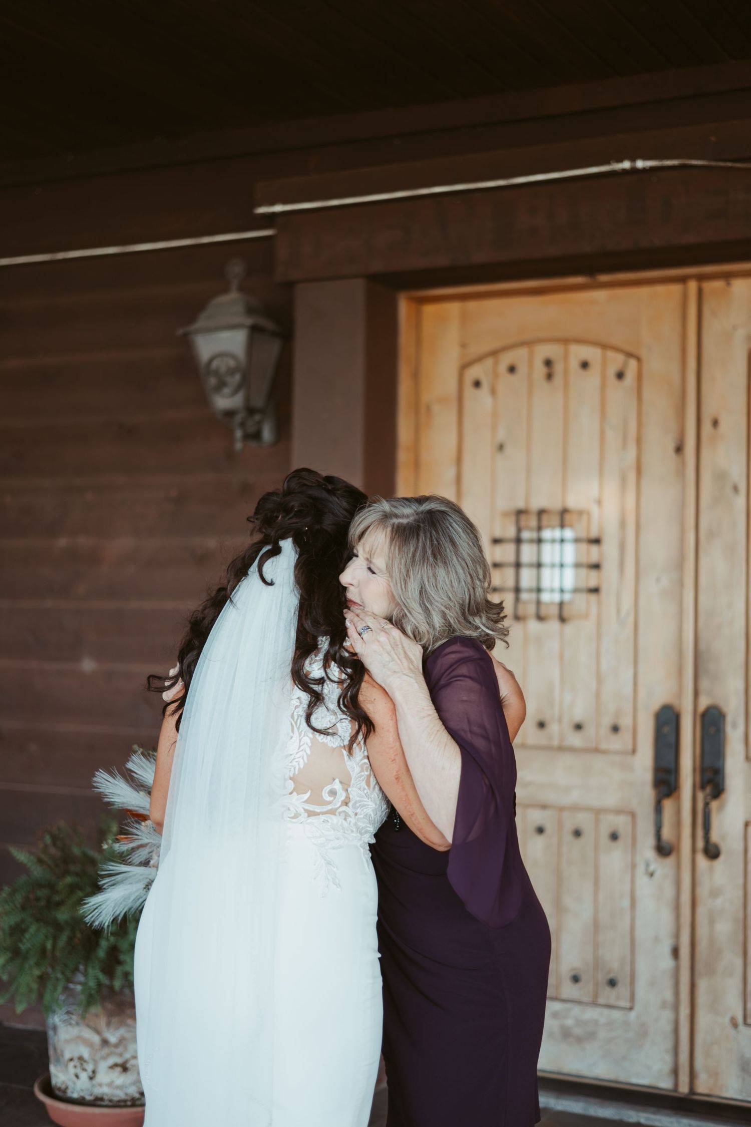 An emotional moment between a bride in a white lace gown and someone in a purple dress at a rustic wooden venue. Wedding photographer DFW