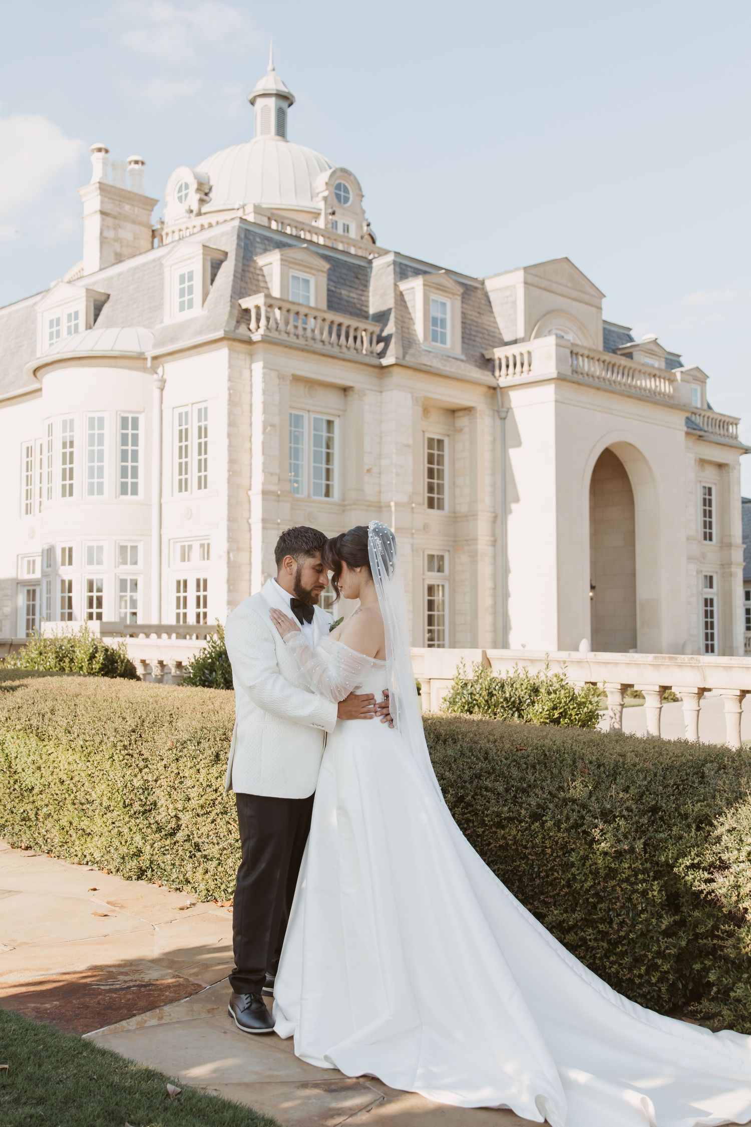 Wedding couple embraces in front of grand white mansion with French architectural details.