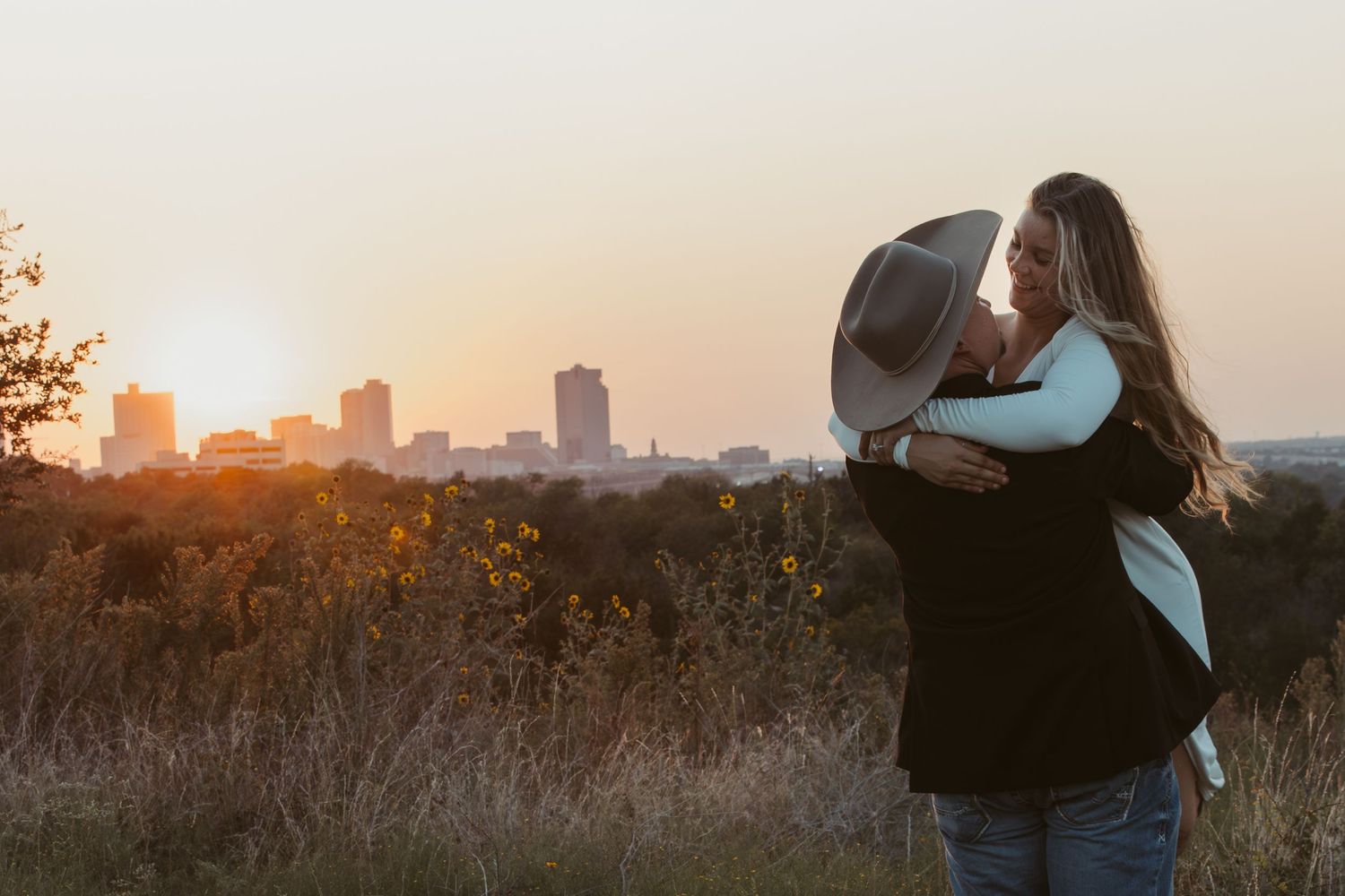 Couple embraces in silhouette against sunset sky with tall city buildings visible in the background.
