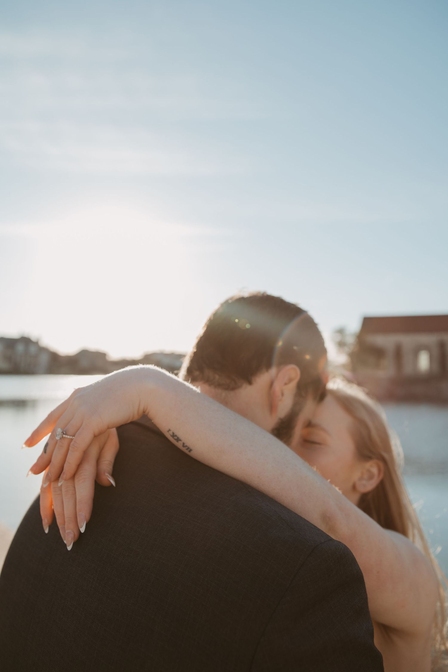 A couple shares an intimate embrace during golden hour by the waterfront as sunlight creates a warm, romantic glow.