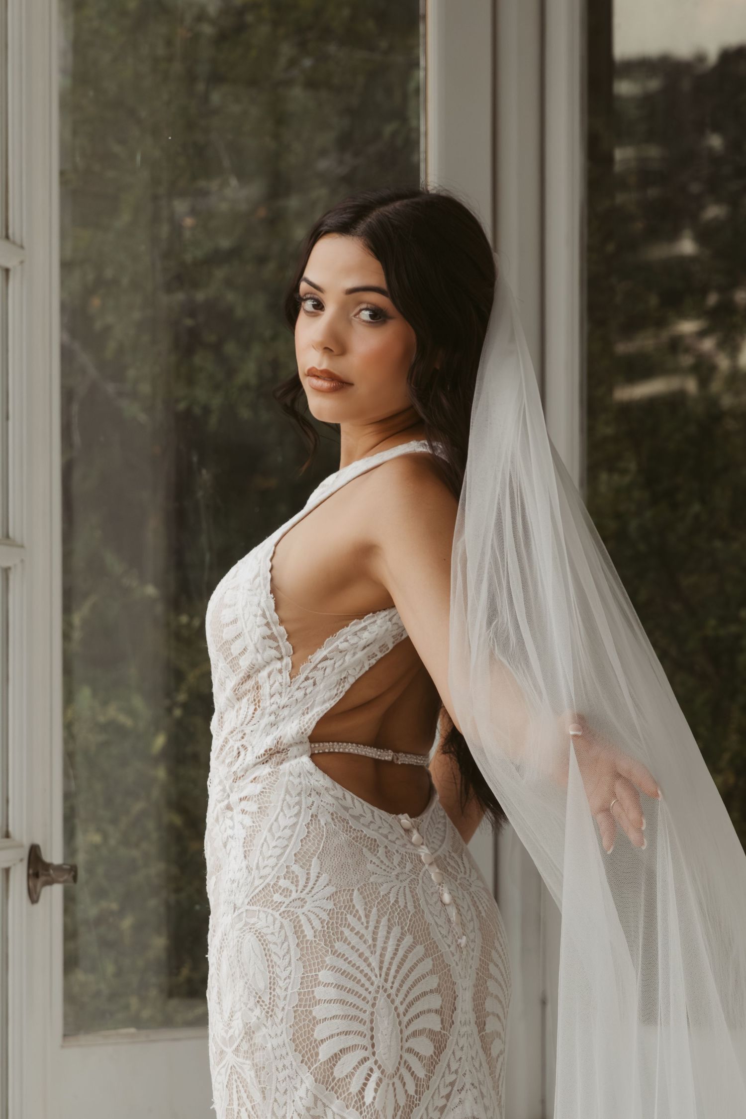 Elegant lace wedding dress with intricate embroidery details shown in a bridal portrait against white curtains.