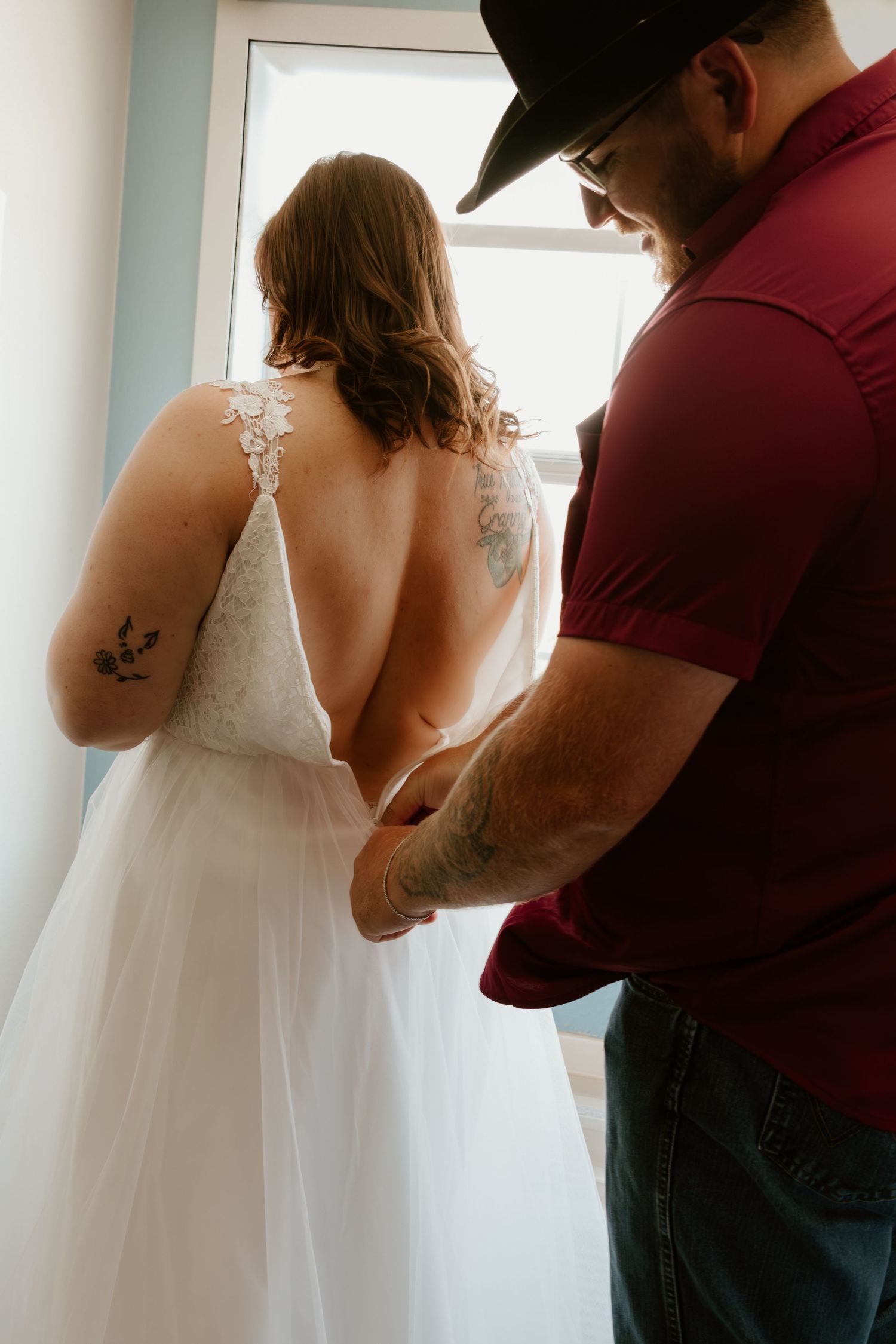 Person in red shirt helping with wedding dress preparations near window.