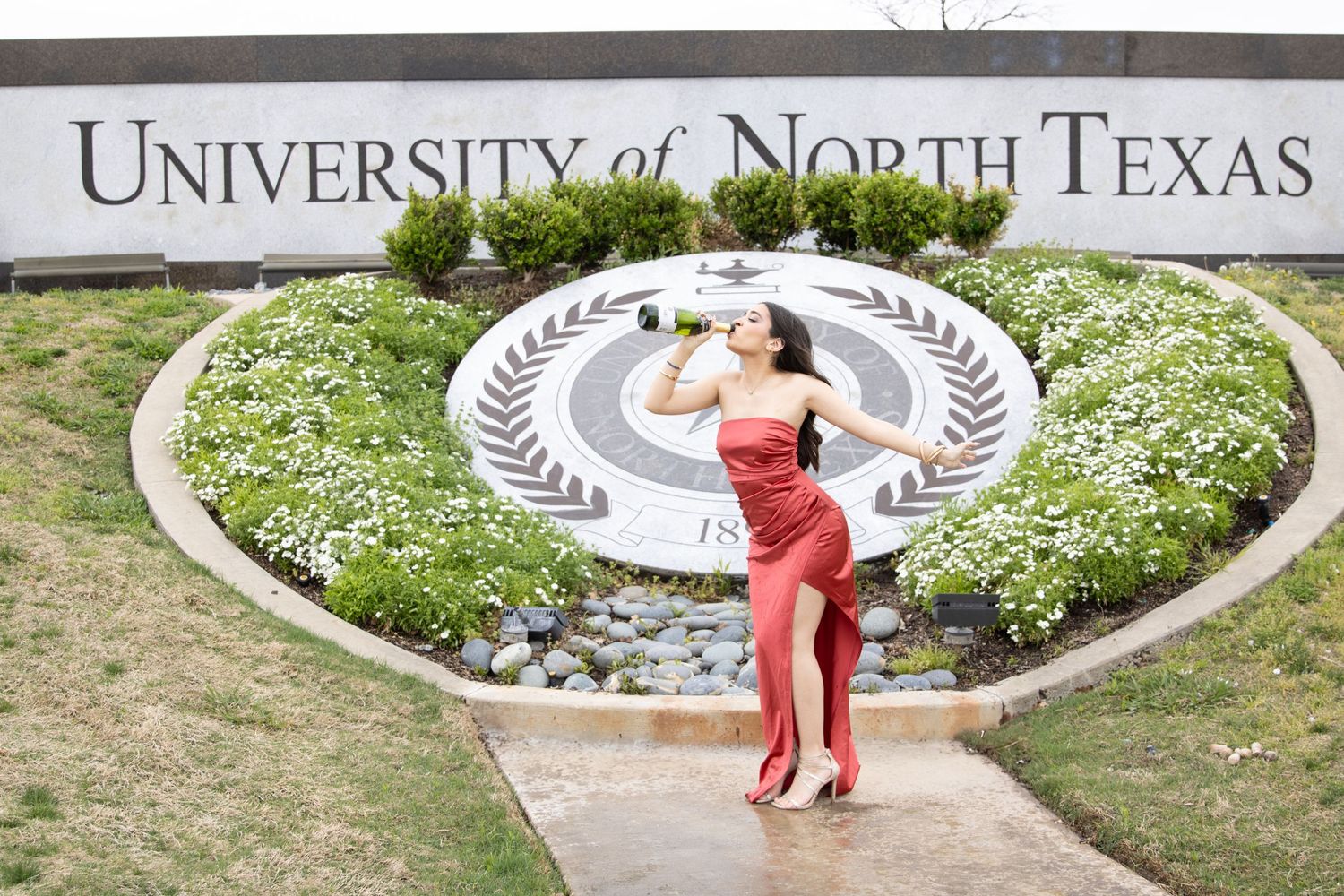 Graduate in red dress poses in front of University of North Texas sign and landscaped garden feature.