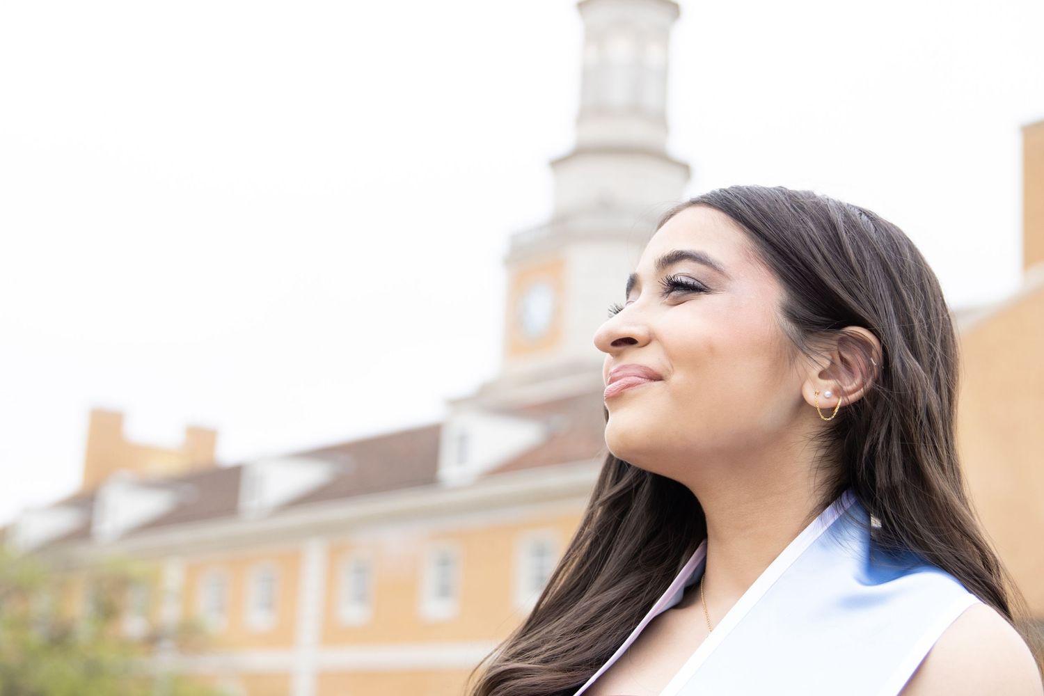 A smiling person with long dark hair wearing a white blazer stands in front of a yellow building with a white spire.