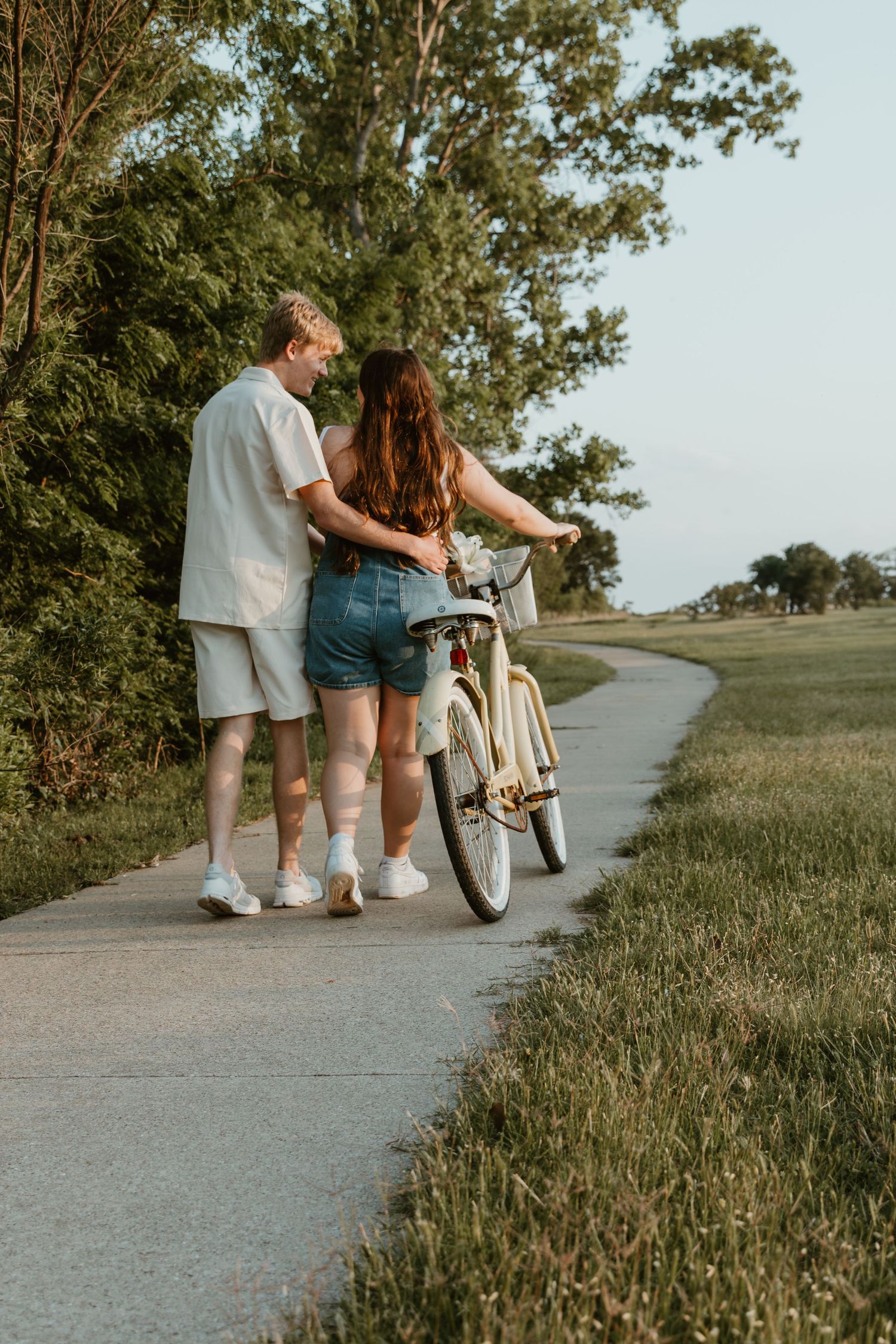 A couple walking along a path on a summer day with a white bicycle beside them in a park-like setting.