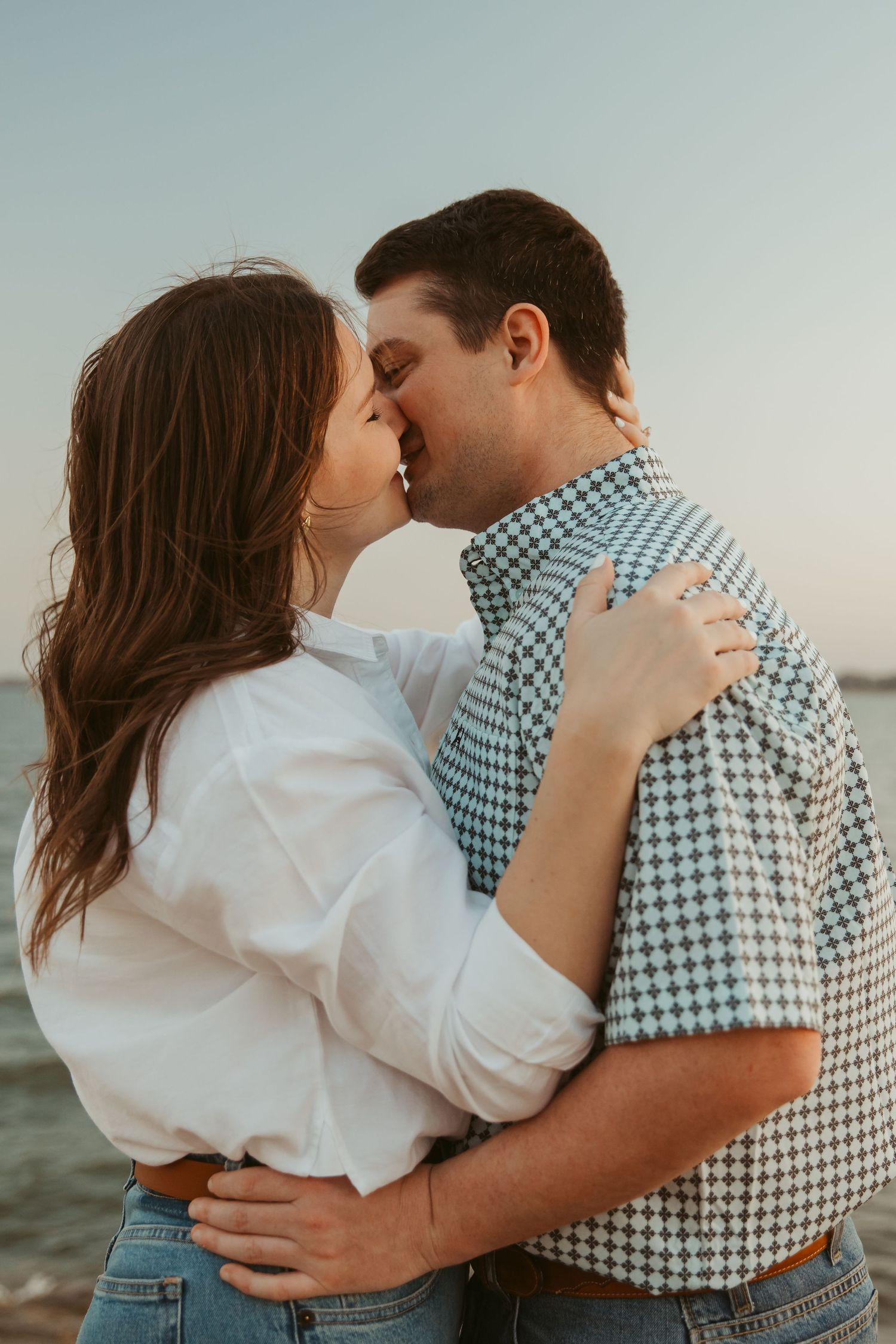 Couple sharing a tender kiss outdoors near water at golden hour, woman wearing engagement ring.