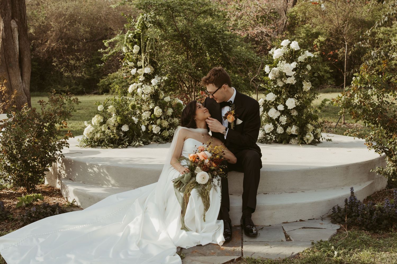 Bride and groom share a romantic kiss on a white stone bench surrounded by lush floral arrangements in an outdoor garden setting.