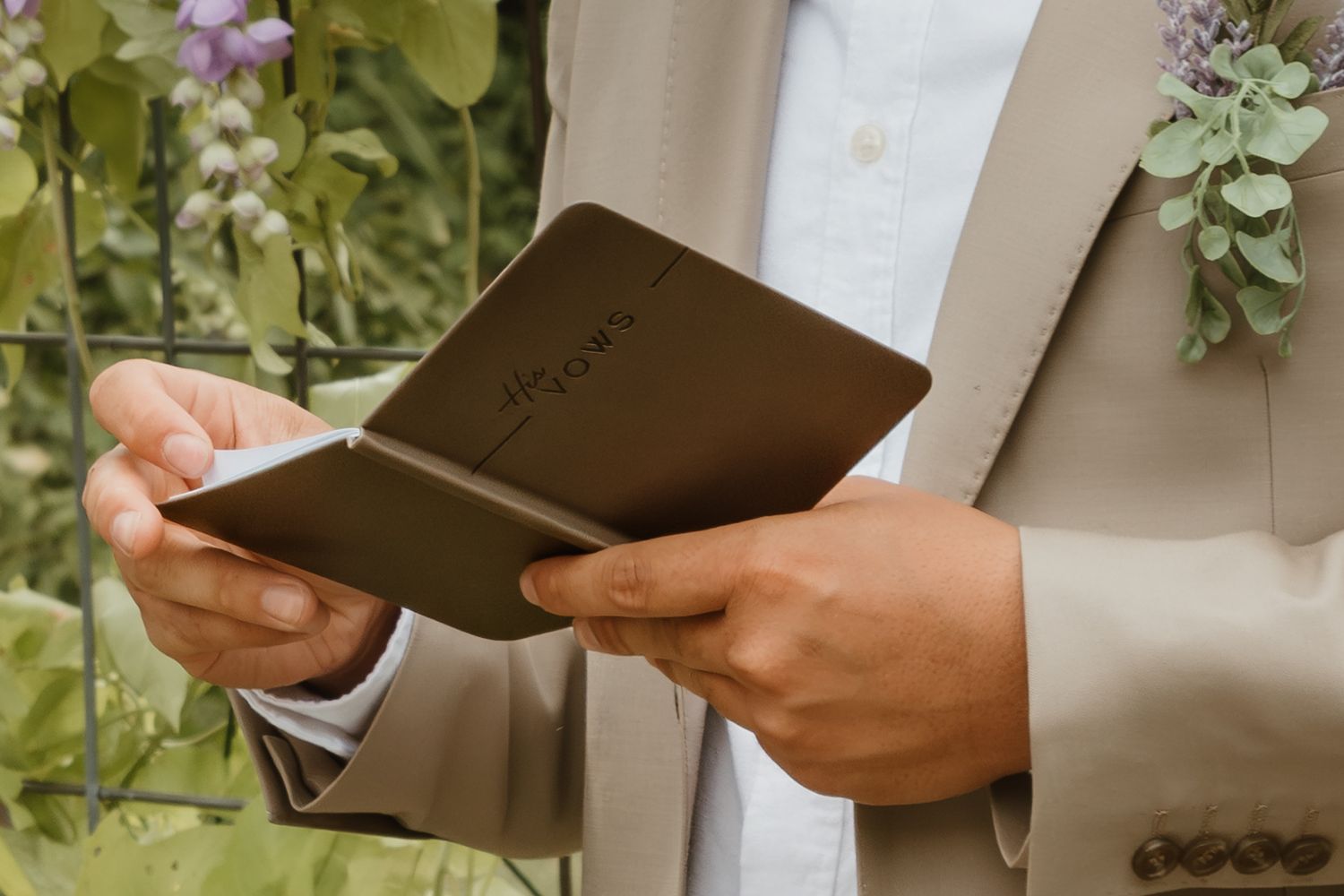Hands in formal attire holding open a brown leather journal against a garden background.