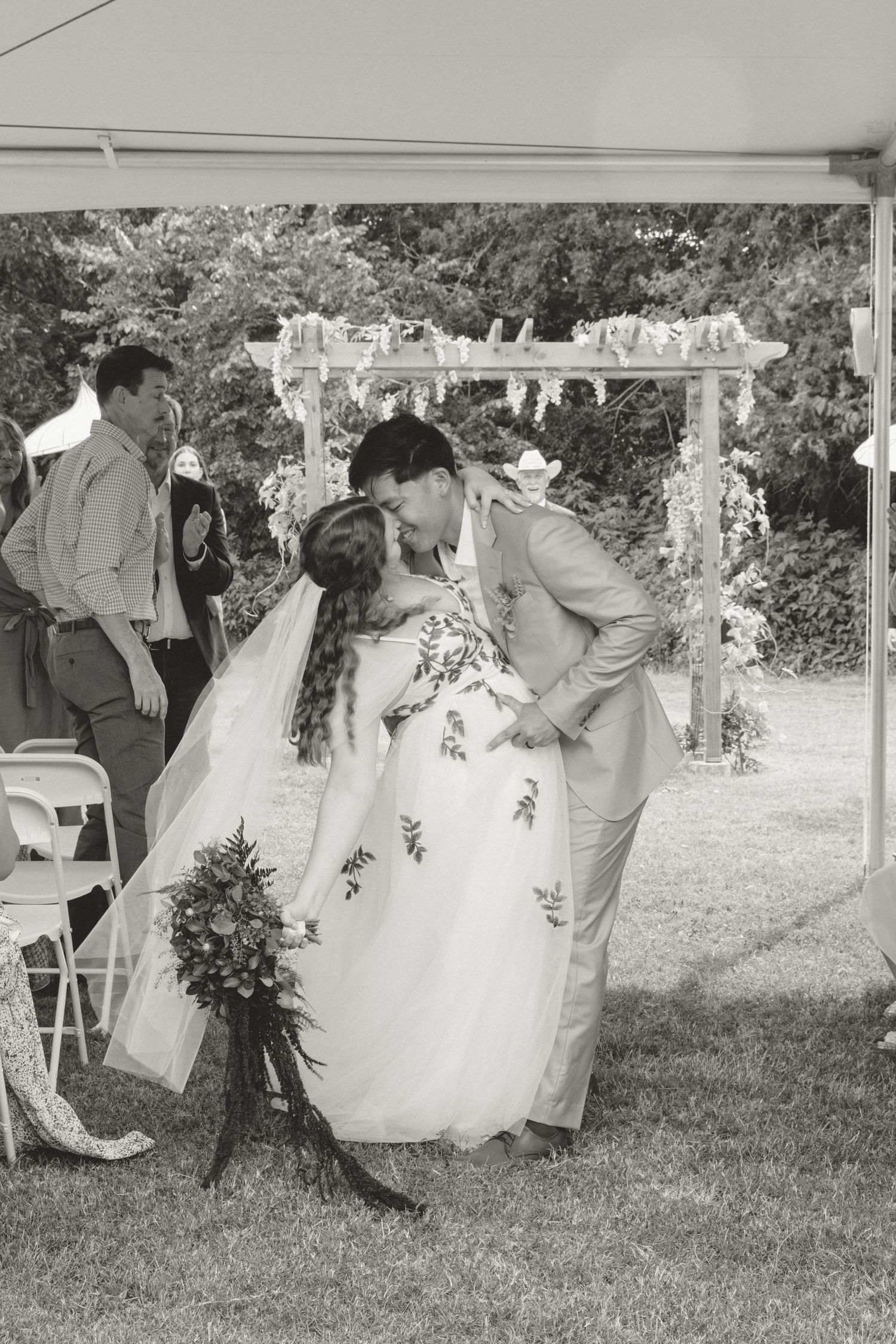 A romantic moment under a floral arch at an outdoor wedding ceremony, captured in black and white photography.