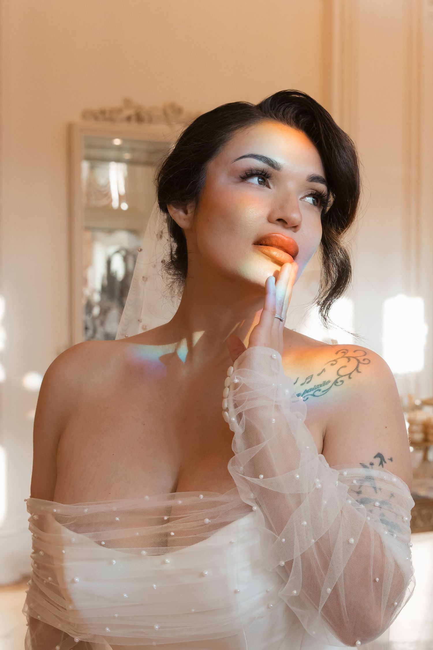 Artistic bridal portrait in soft lighting showing sheer tulle sleeves, delicate tattoo details and glowing orange lipstick.
