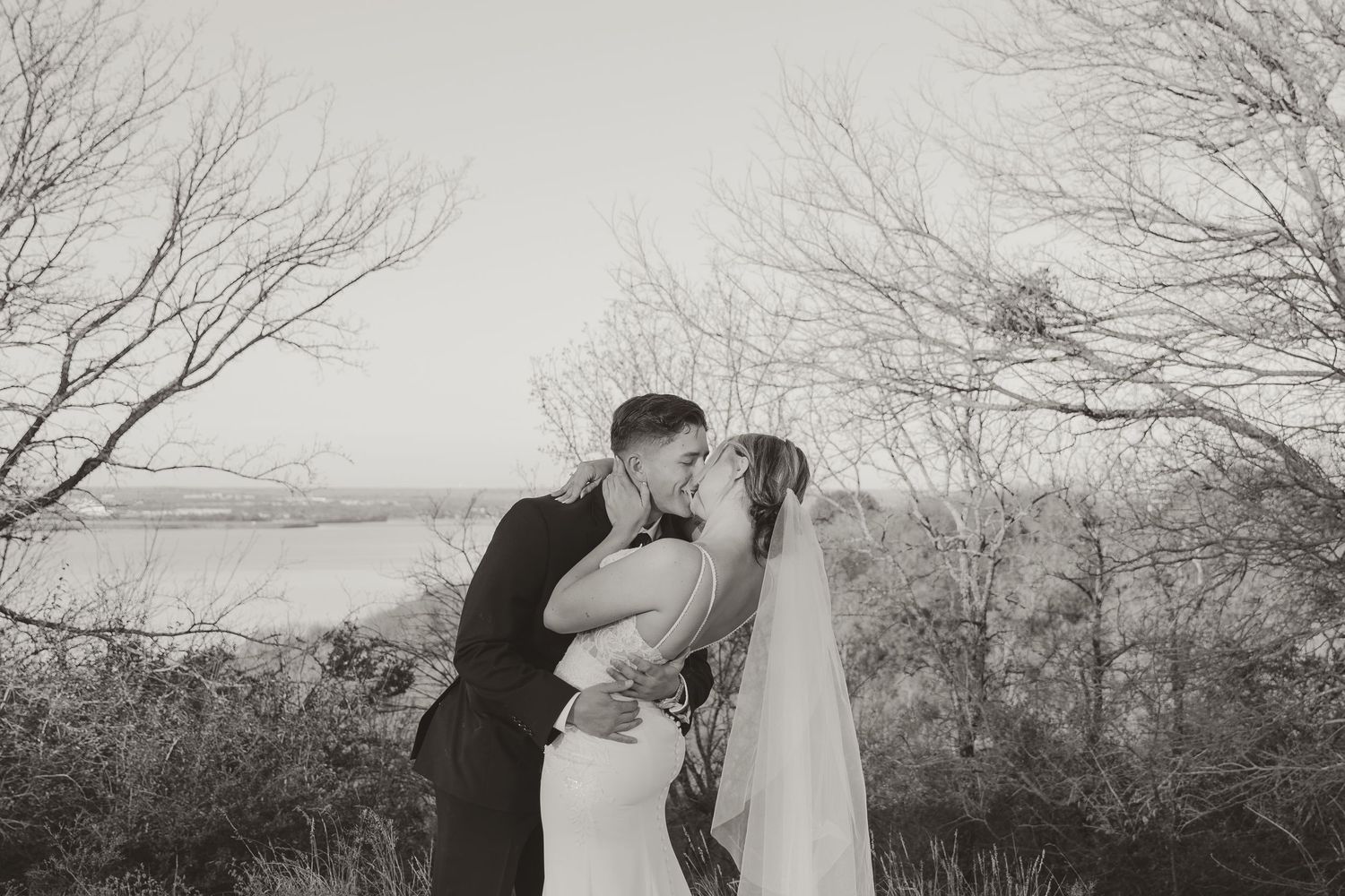 A romantic black and white wedding photograph taken outdoors with bare trees and water in the background.