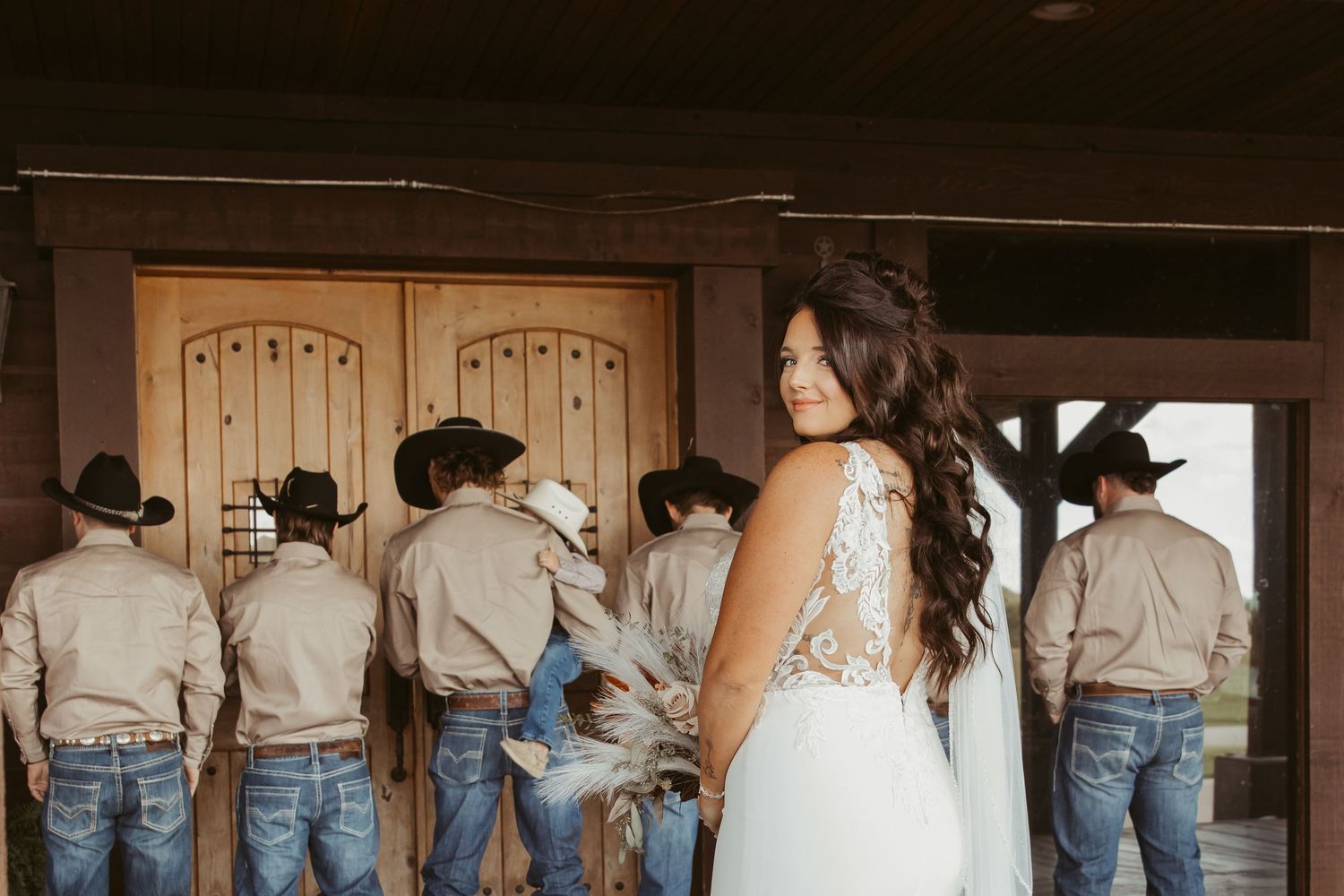 A bride in a white lace dress stands near a group of groomsmen wearing cowboy hats and jeans at a rustic venue. Wedding photographer DFW