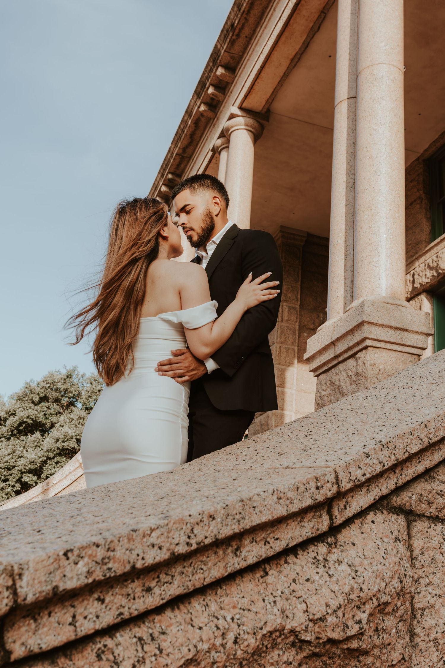 A romantic couple embraces against a backdrop of classical stone columns in warm sunlight.