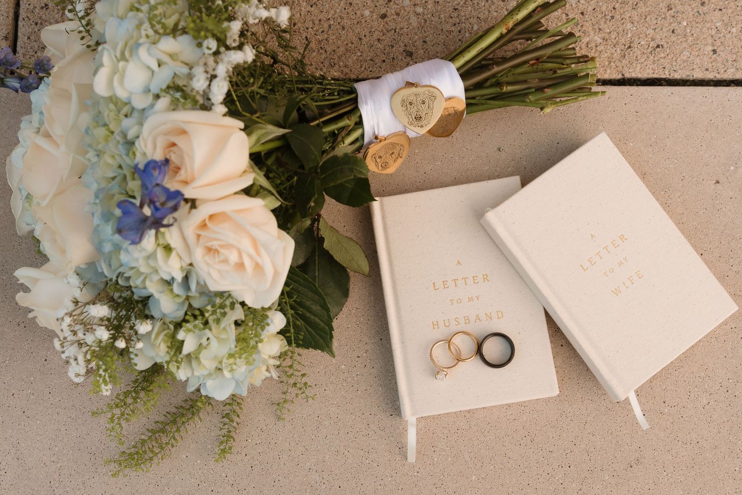 A delicate floral arrangement of white roses and blue hydrangeas with wedding rings on a cream-colored surface. Wedding Photographer DFW