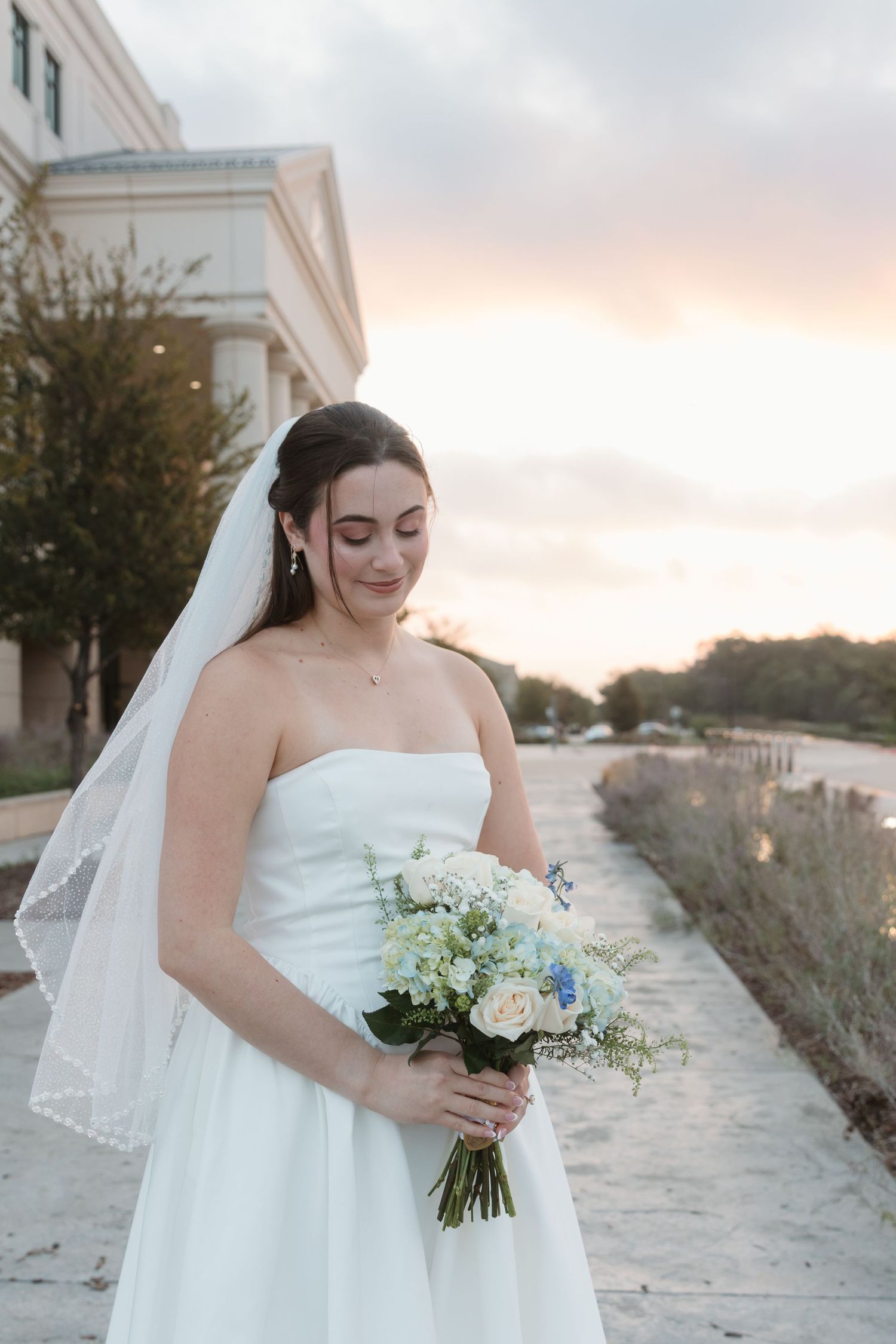 A bride in a white strapless gown holds a delicate bouquet of white flowers at sunset.