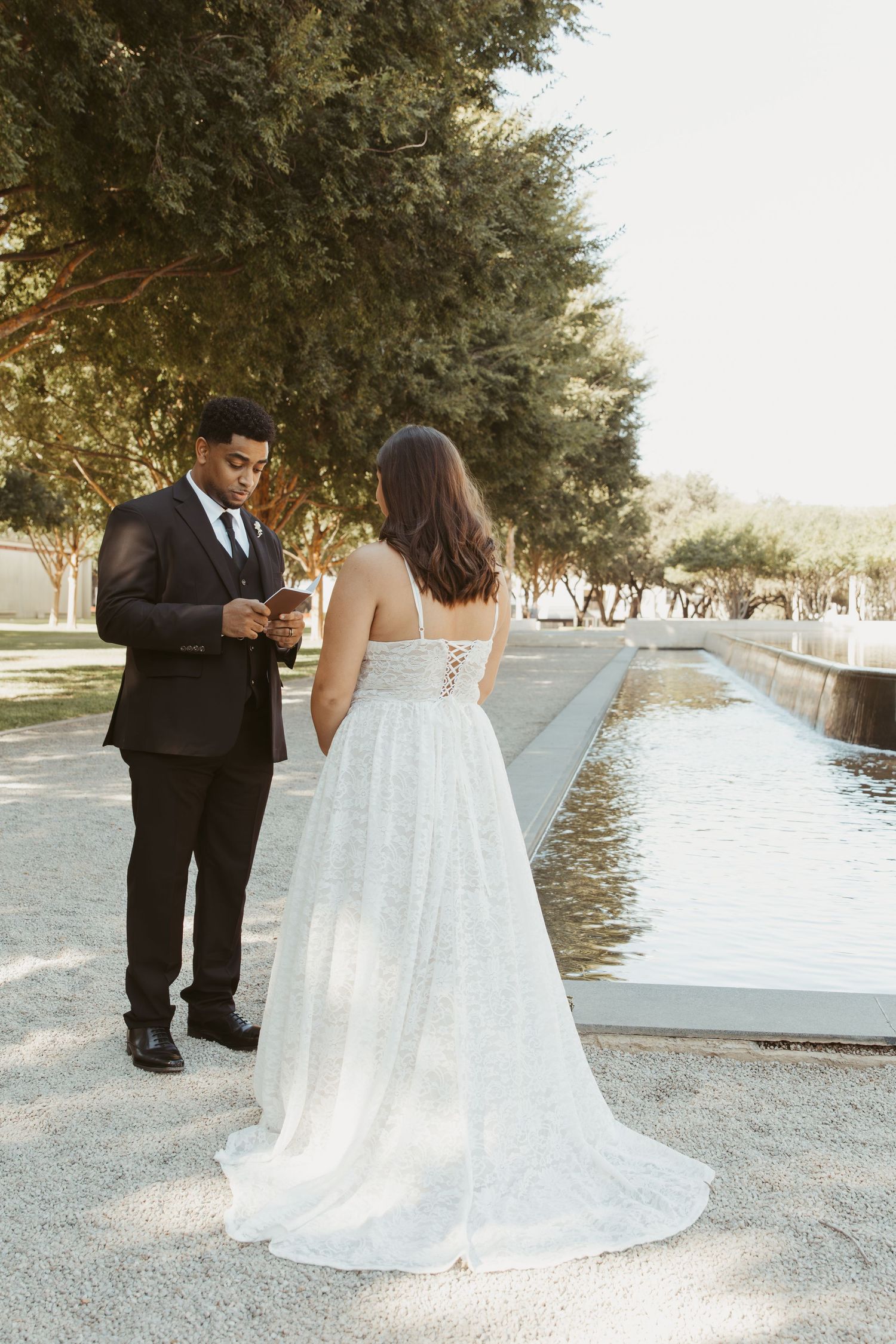 A couple in wedding attire stands by a reflecting pool in a garden setting during golden hour. Wedding Photographer DFW