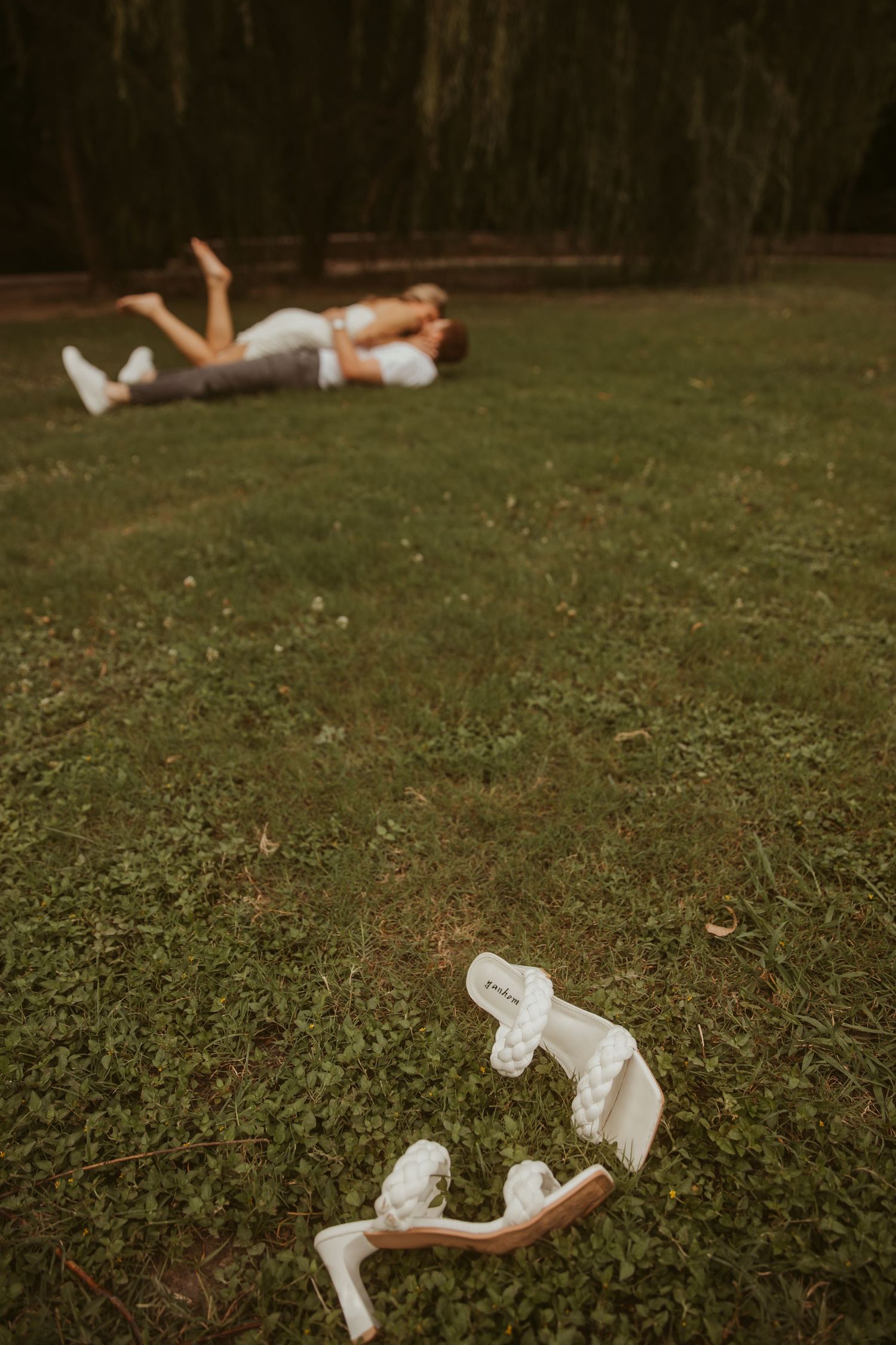 Couple lying on green grass in a park, with white bridal heels discarded in the foreground.