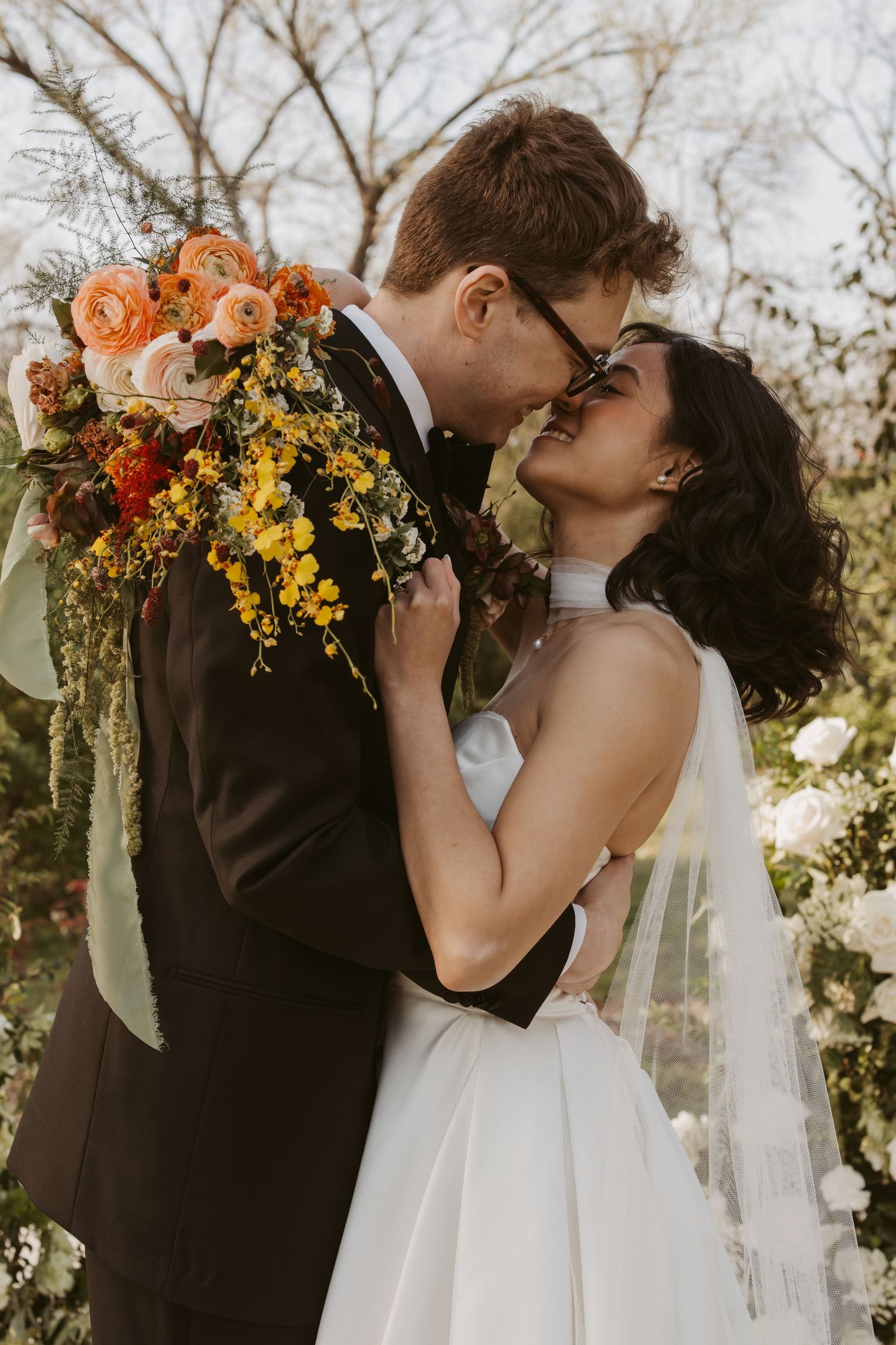 A bride and groom share a romantic moment, holding a vibrant orange and yellow bouquet outdoors.