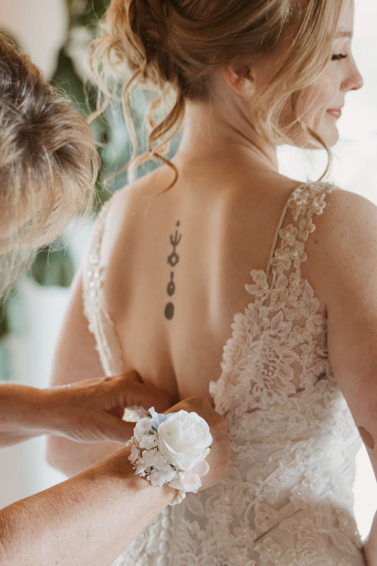 Bride in lace wedding dress with back tattoo gets wrist corsage of white roses fastened by a helper.