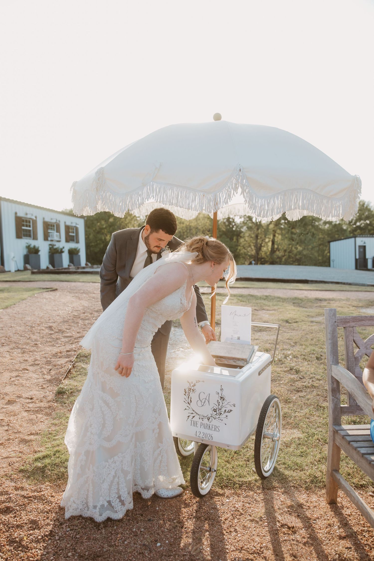 Bride and groom in wedding attire lean over a white ice cream cart with umbrella outdoors at golden hour.