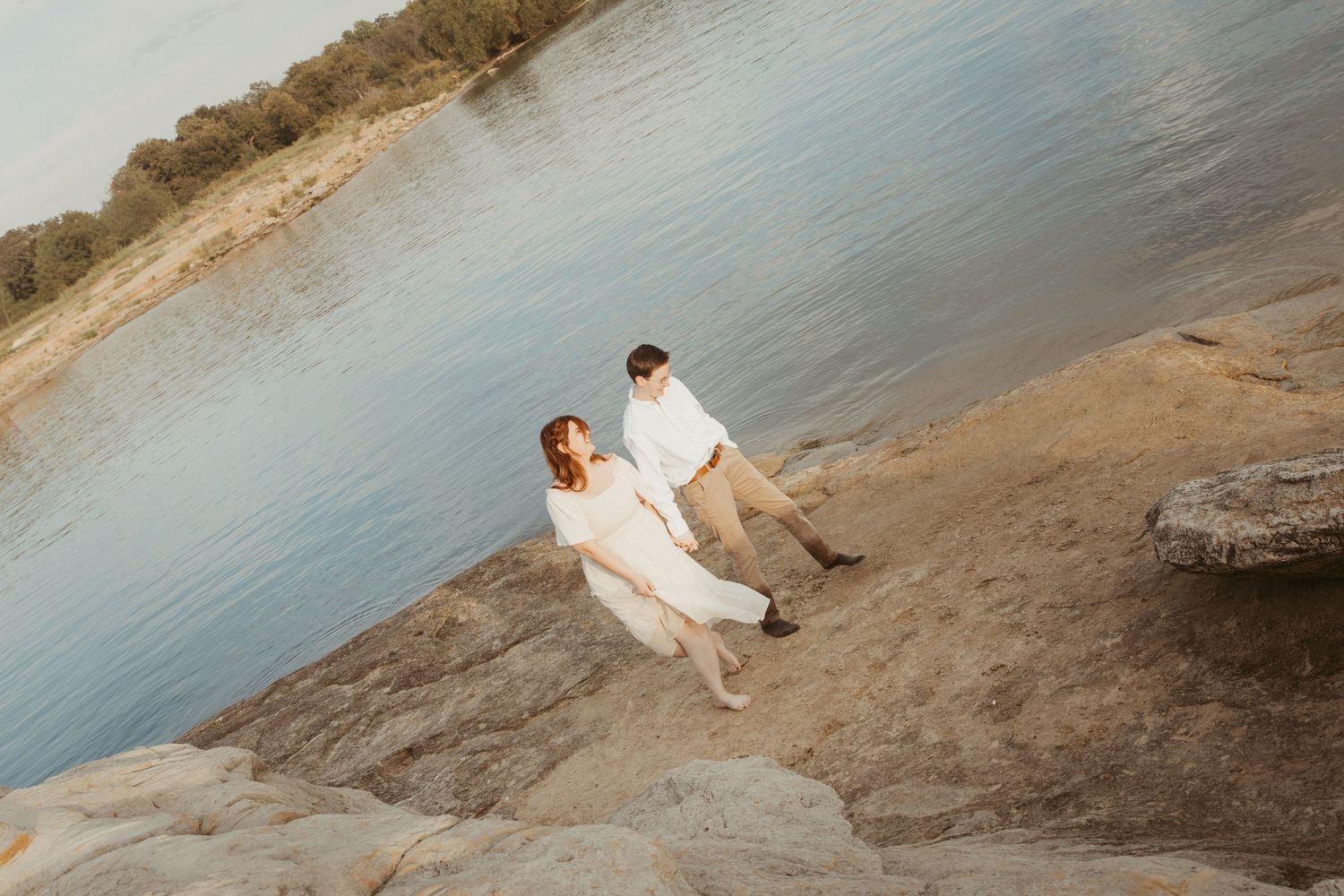 A couple walks along a scenic shoreline with rocky cliffs and calm waters in the background.