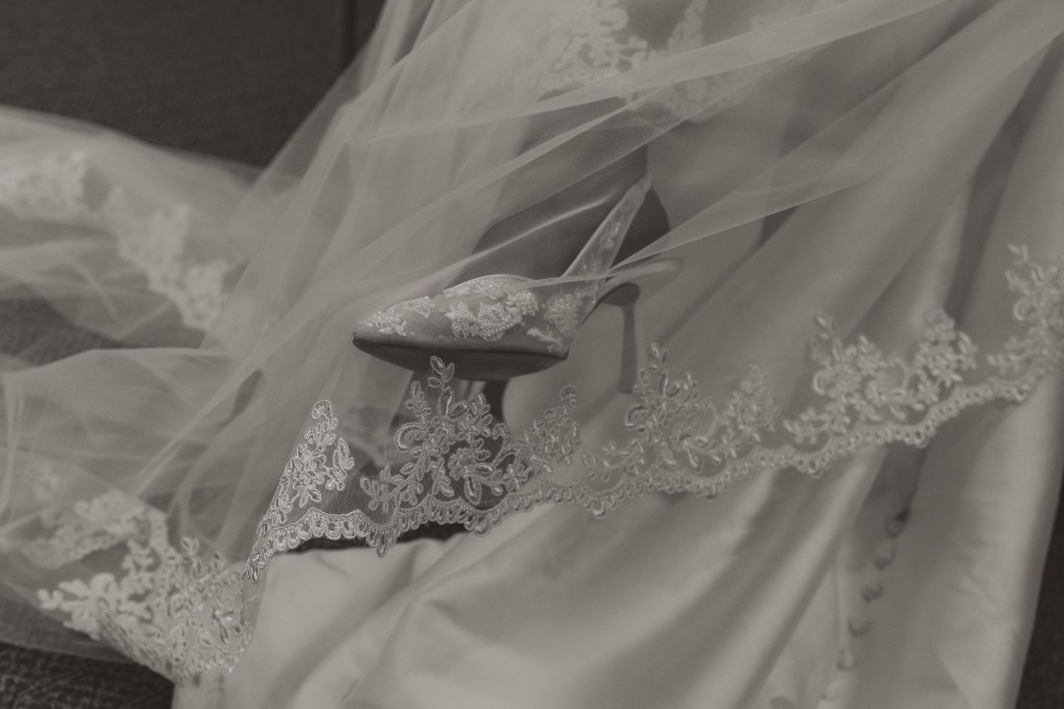 Elegant bridal shoes rest on delicate lace wedding dress and veil in a classic black and white photograph.