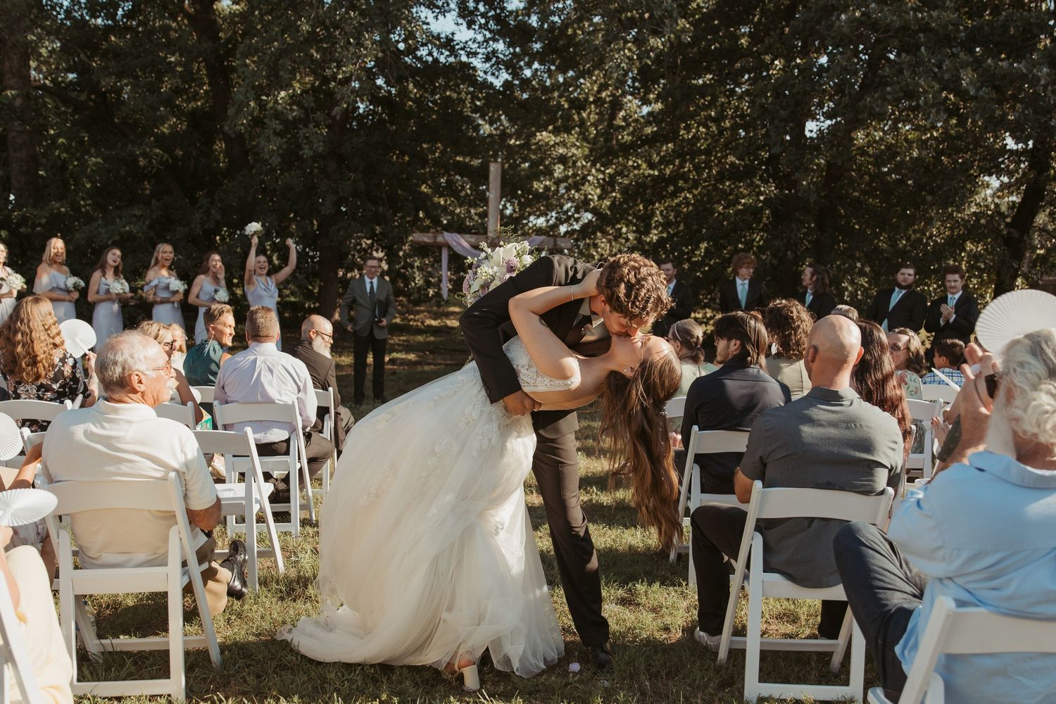 Bride and groom share first kiss at outdoor wedding ceremony with guests seated on white chairs under trees.