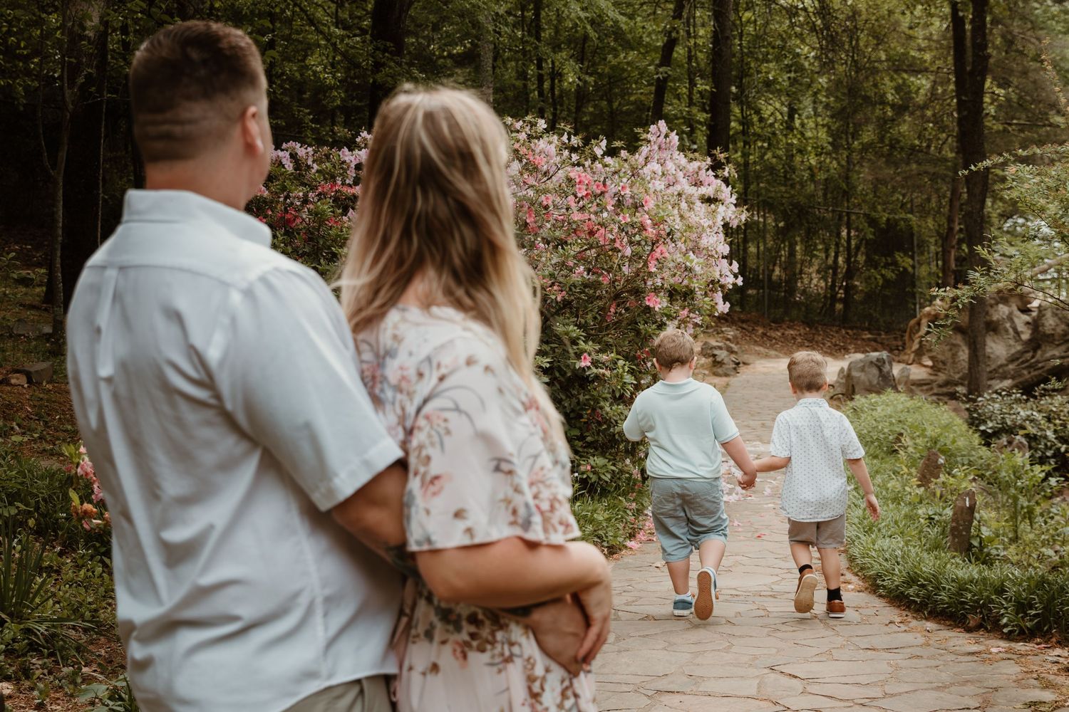 Two pairs of people walk along a garden path surrounded by blooming flowers and trees.