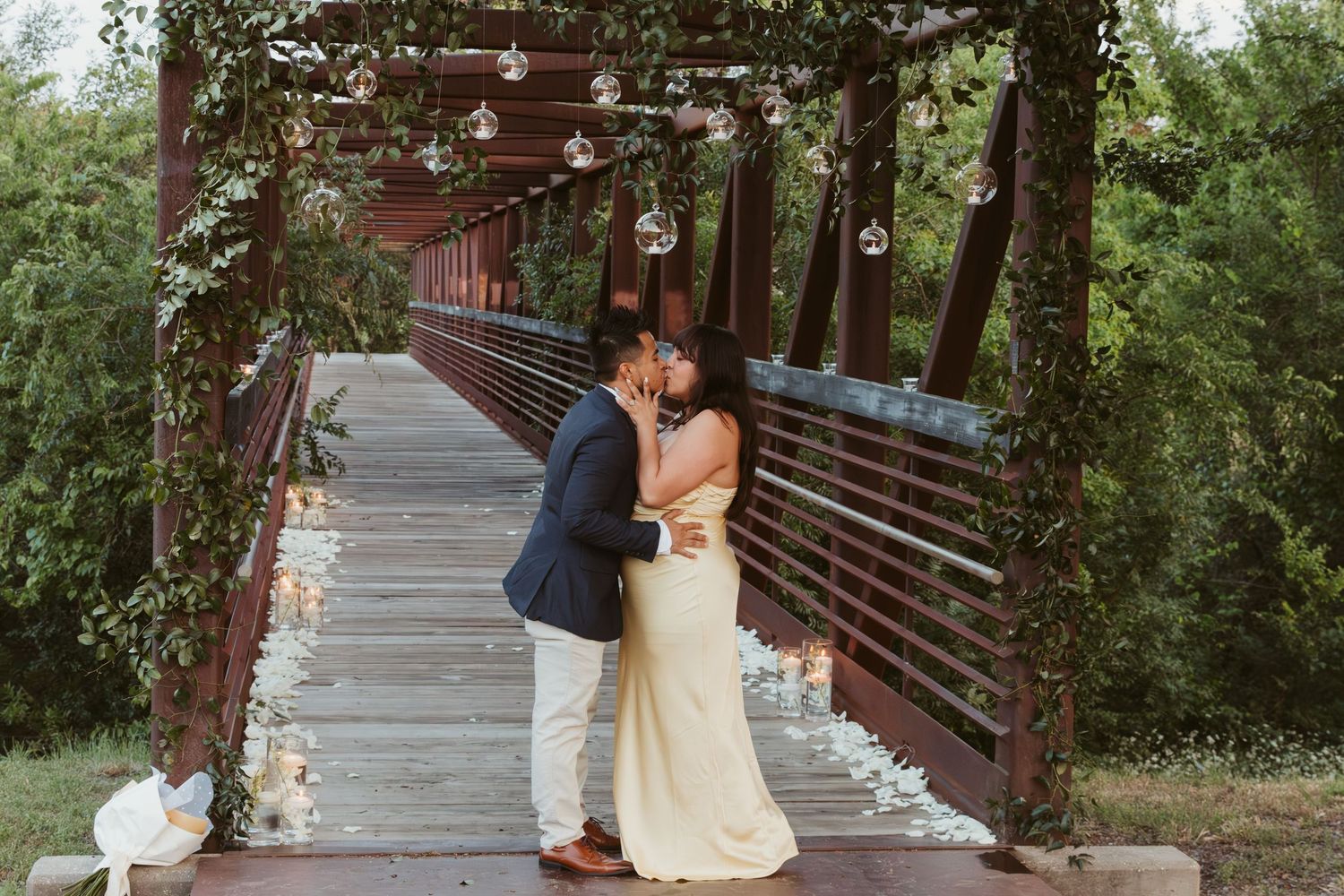 Beautiful wedding couple embraces on wooden bridge decorated with string lights and greenery.