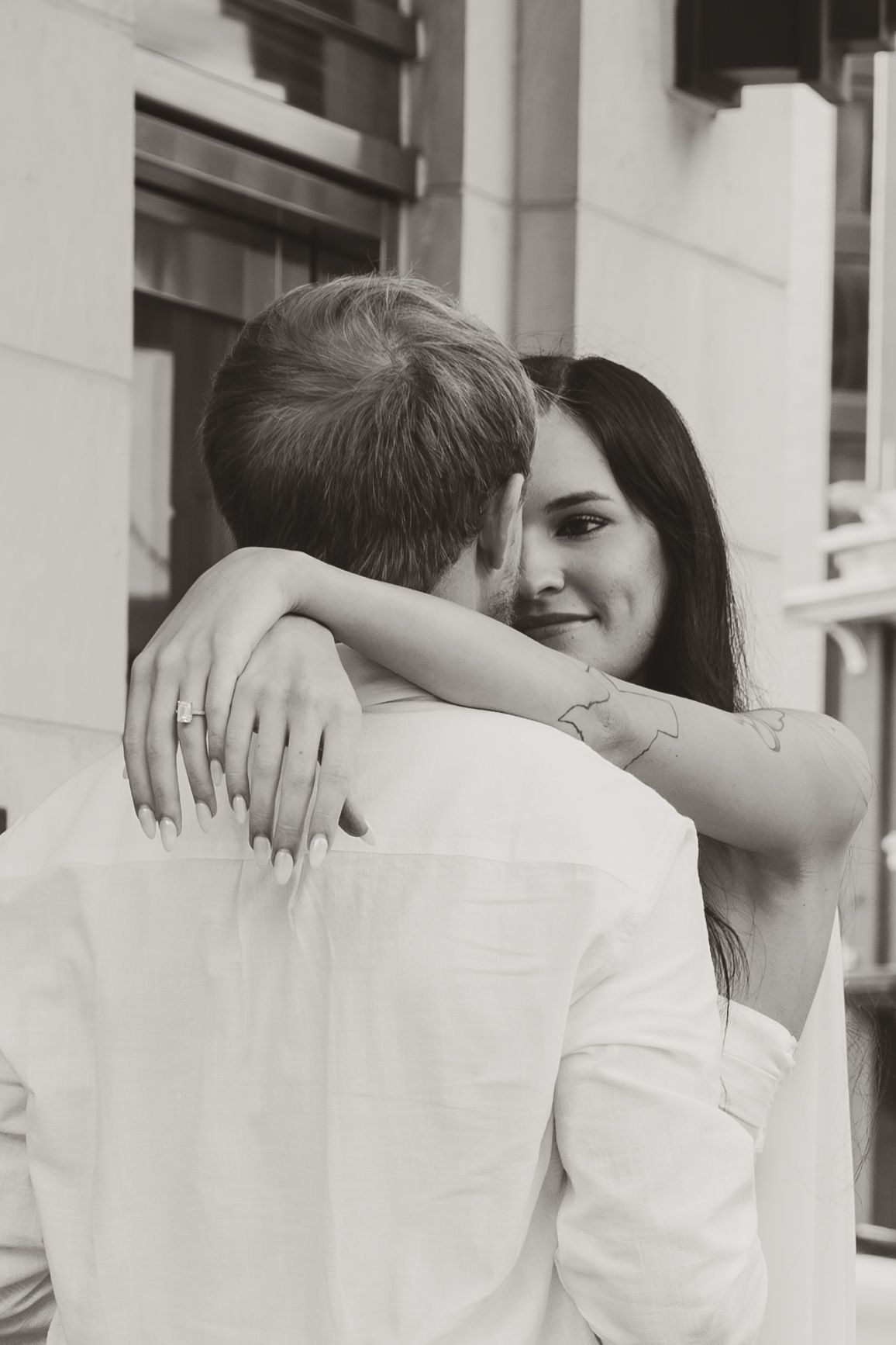 A romantic black and white photo of a couple embracing, with a smiling woman looking over her partner's shoulder.