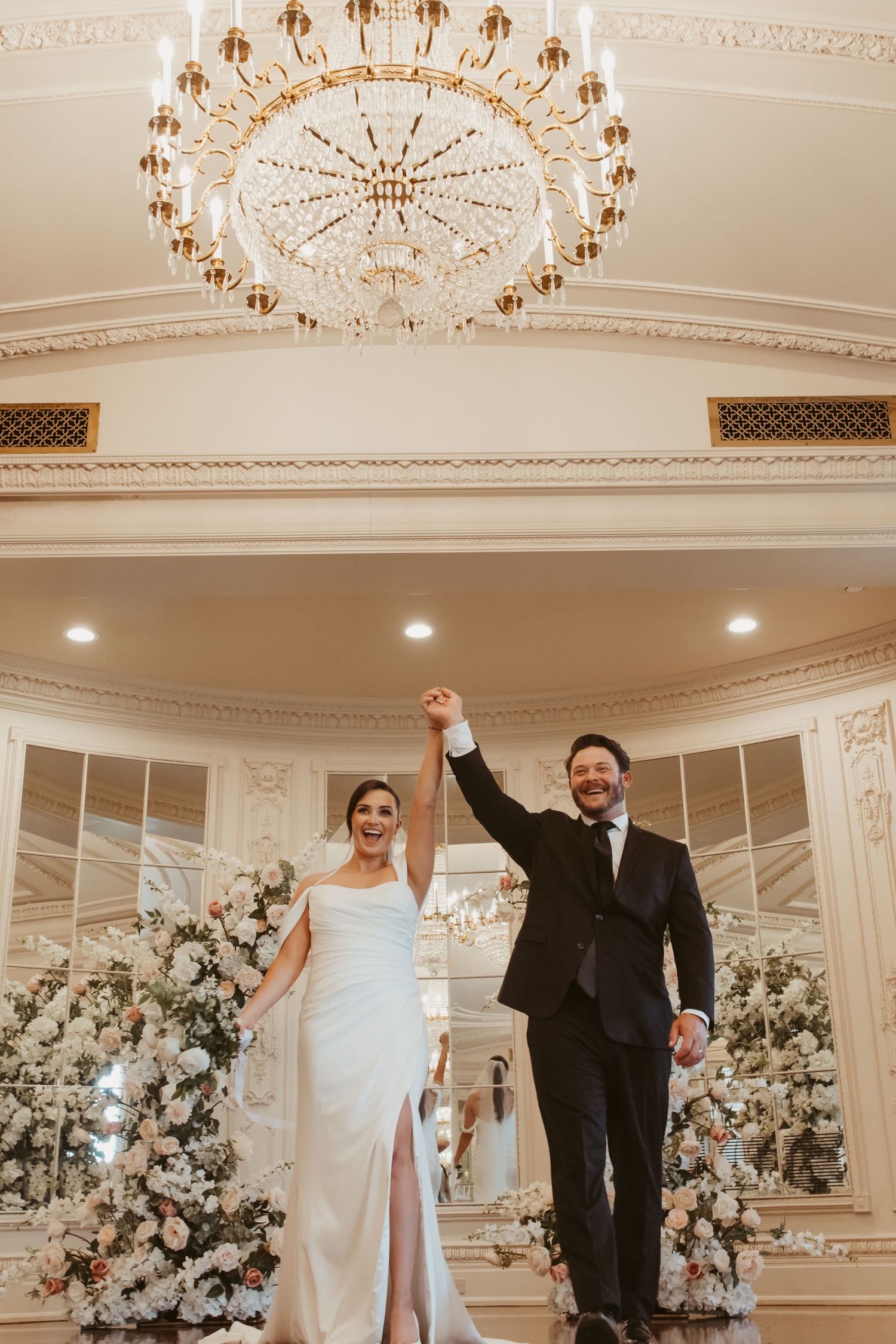 Newlyweds celebrate under crystal chandeliers in luxurious white ballroom with floral arrangements.