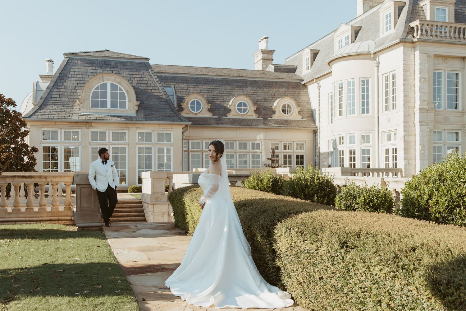 Elegant wedding couple stands on terrace of luxurious French-style mansion with ornate architecture and manicured hedges.