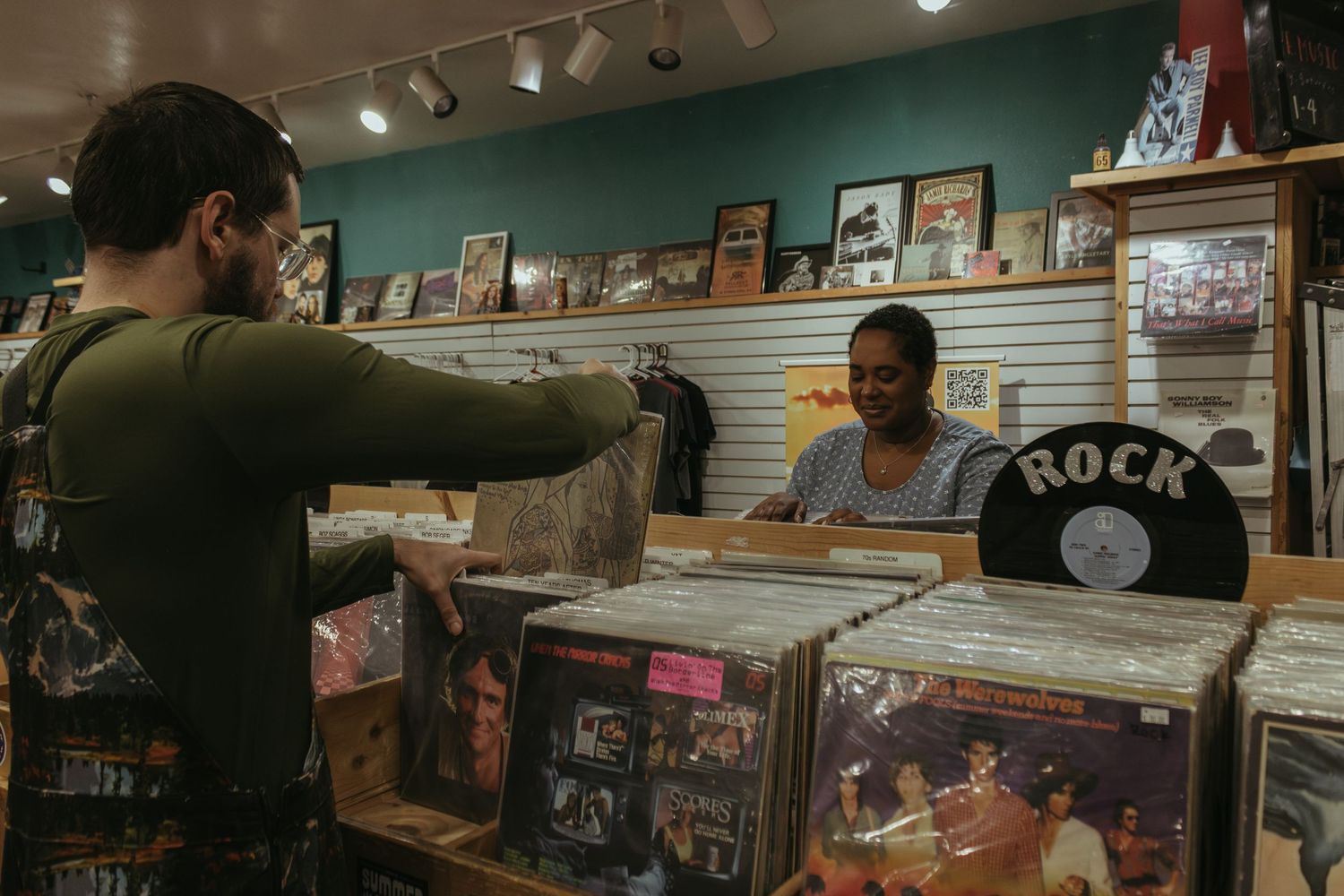Two people browsing through vinyl records at a music store with framed photos and memorabilia on the teal wall.
