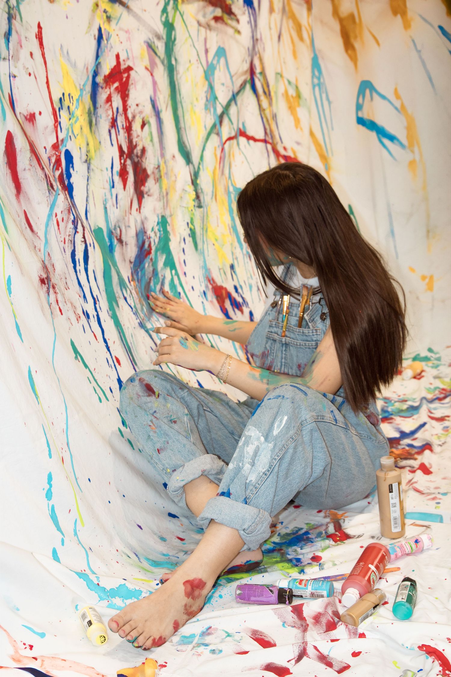 Someone in overalls sits barefoot on paint-splattered floor creating colorful abstract art on white wall.