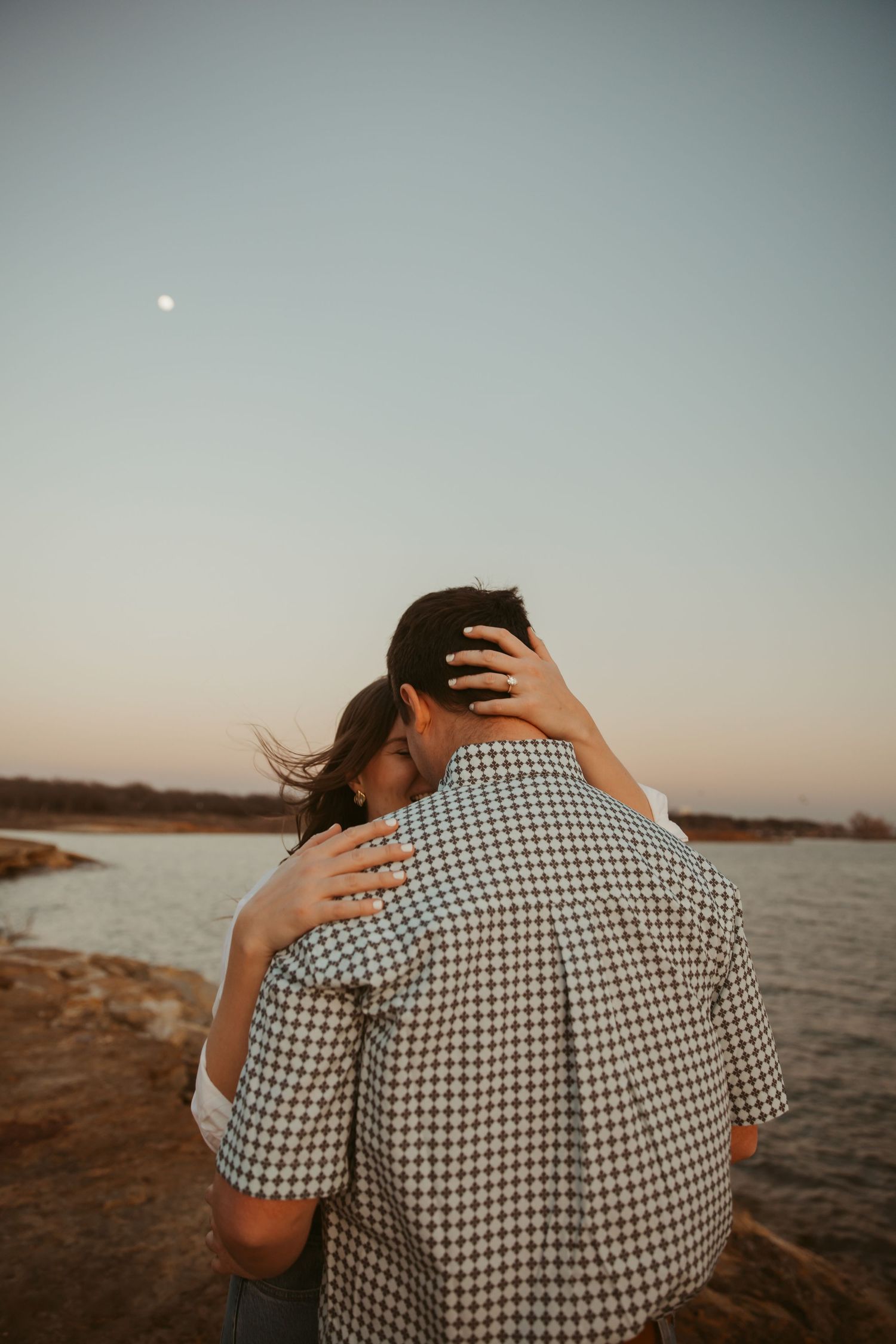 Couple embracing romantically by a lake at dusk with moon visible in soft pastel sky.