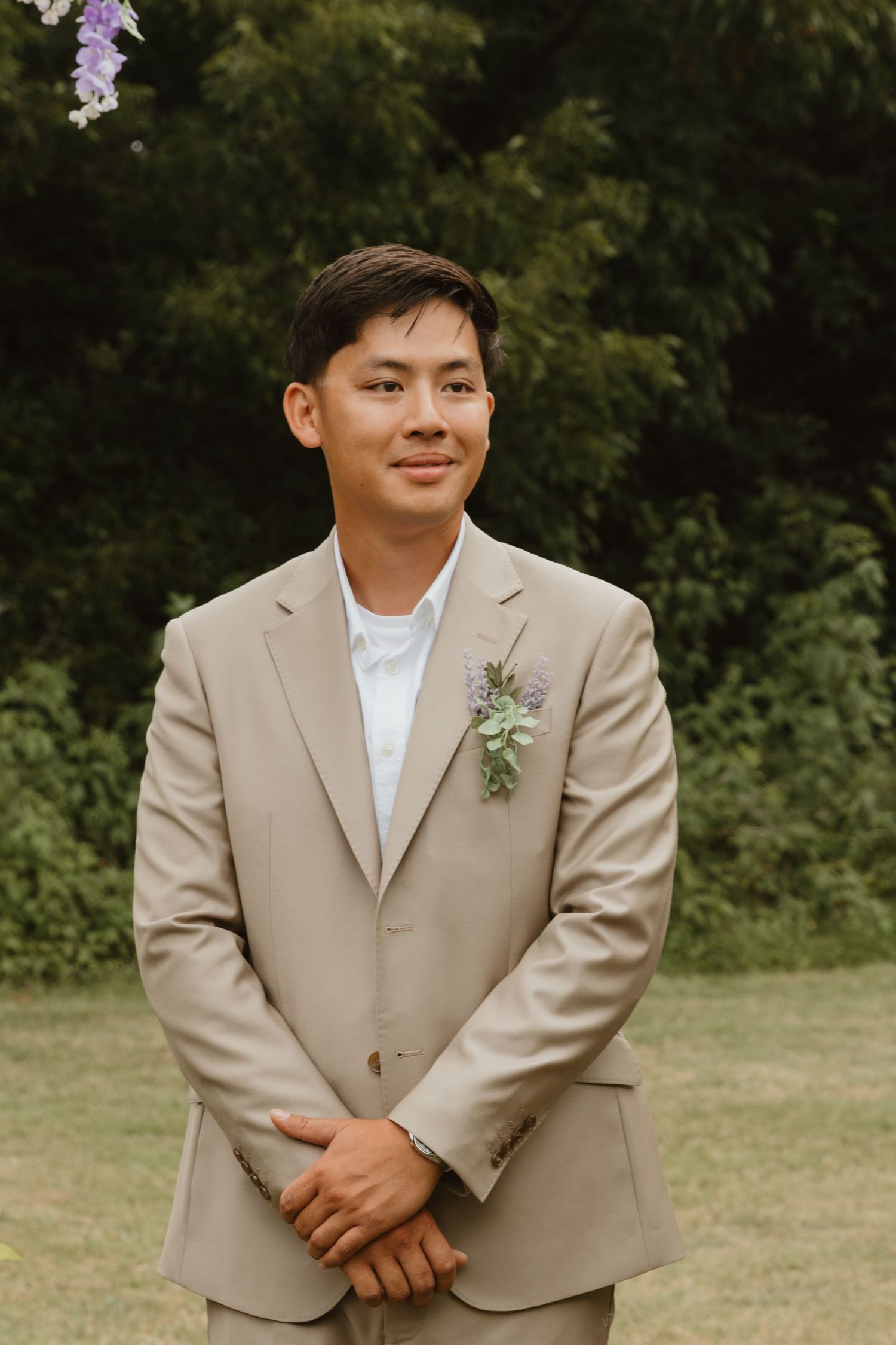 Groomsman in tan suit with boutonniere poses for portraits in an outdoor garden setting.