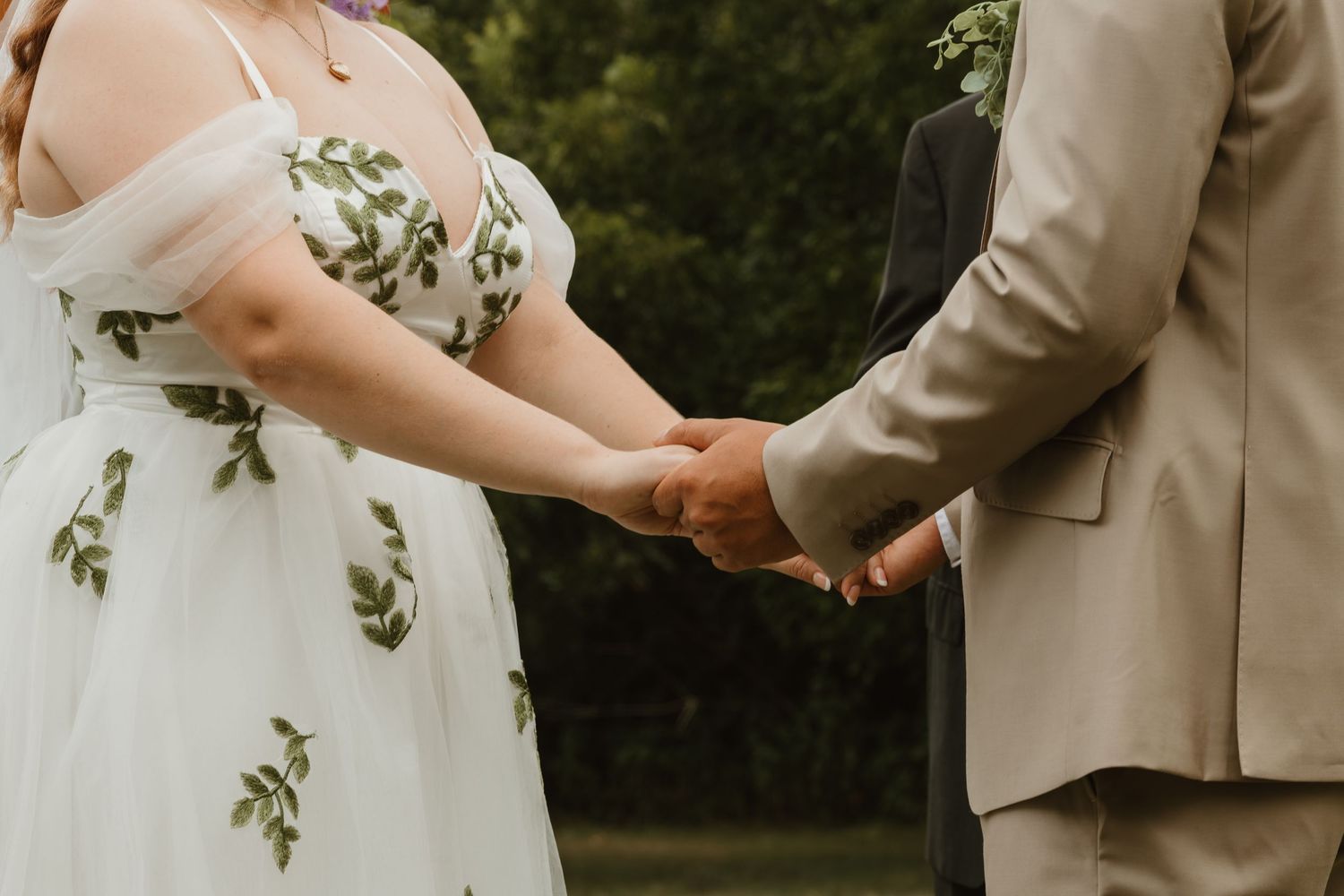 Bride in floral embroidered white gown holds hands with groom in beige suit during outdoor wedding ceremony.