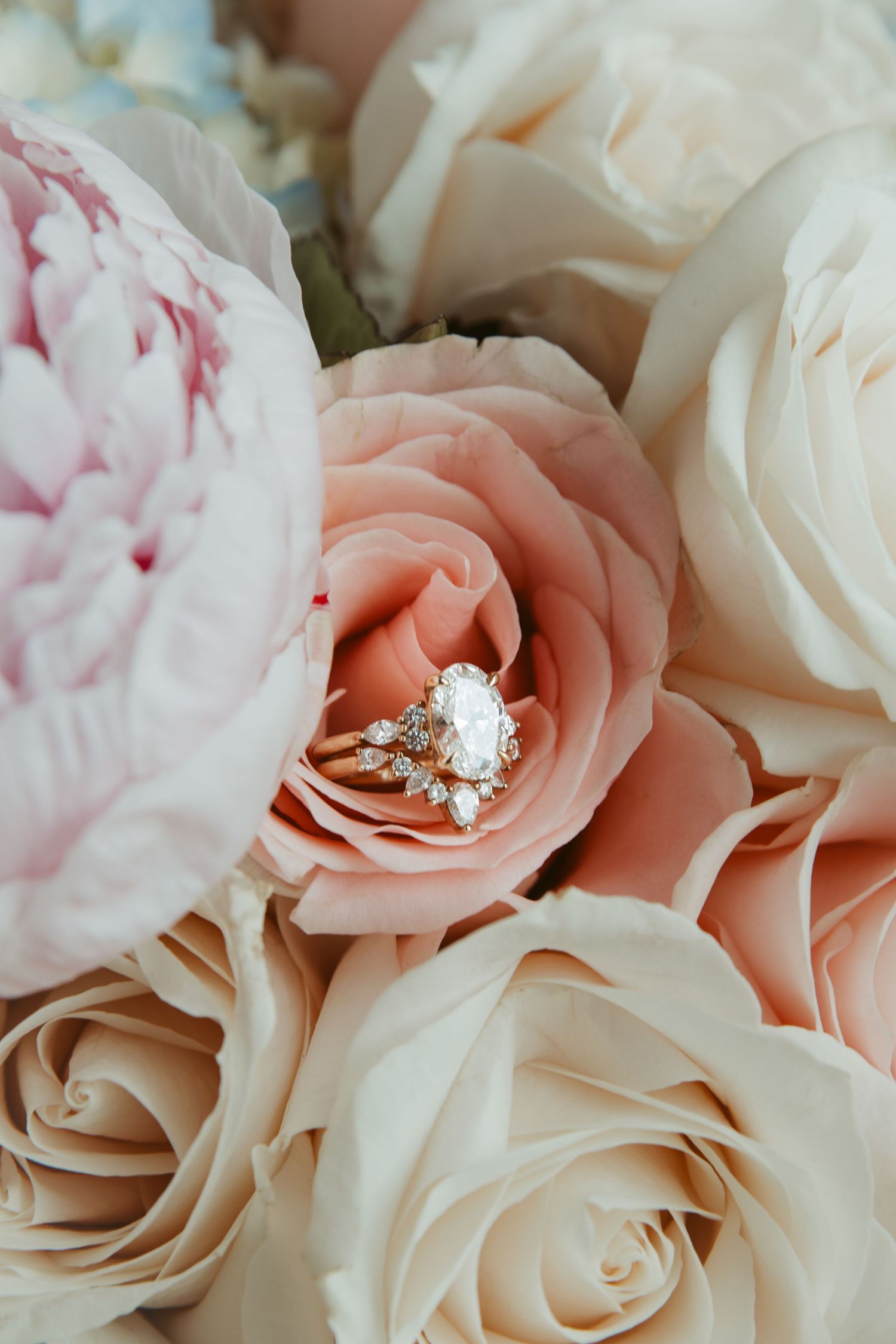 A beautiful diamond engagement ring nestled among soft pink and white roses and peonies in a romantic floral arrangement.
