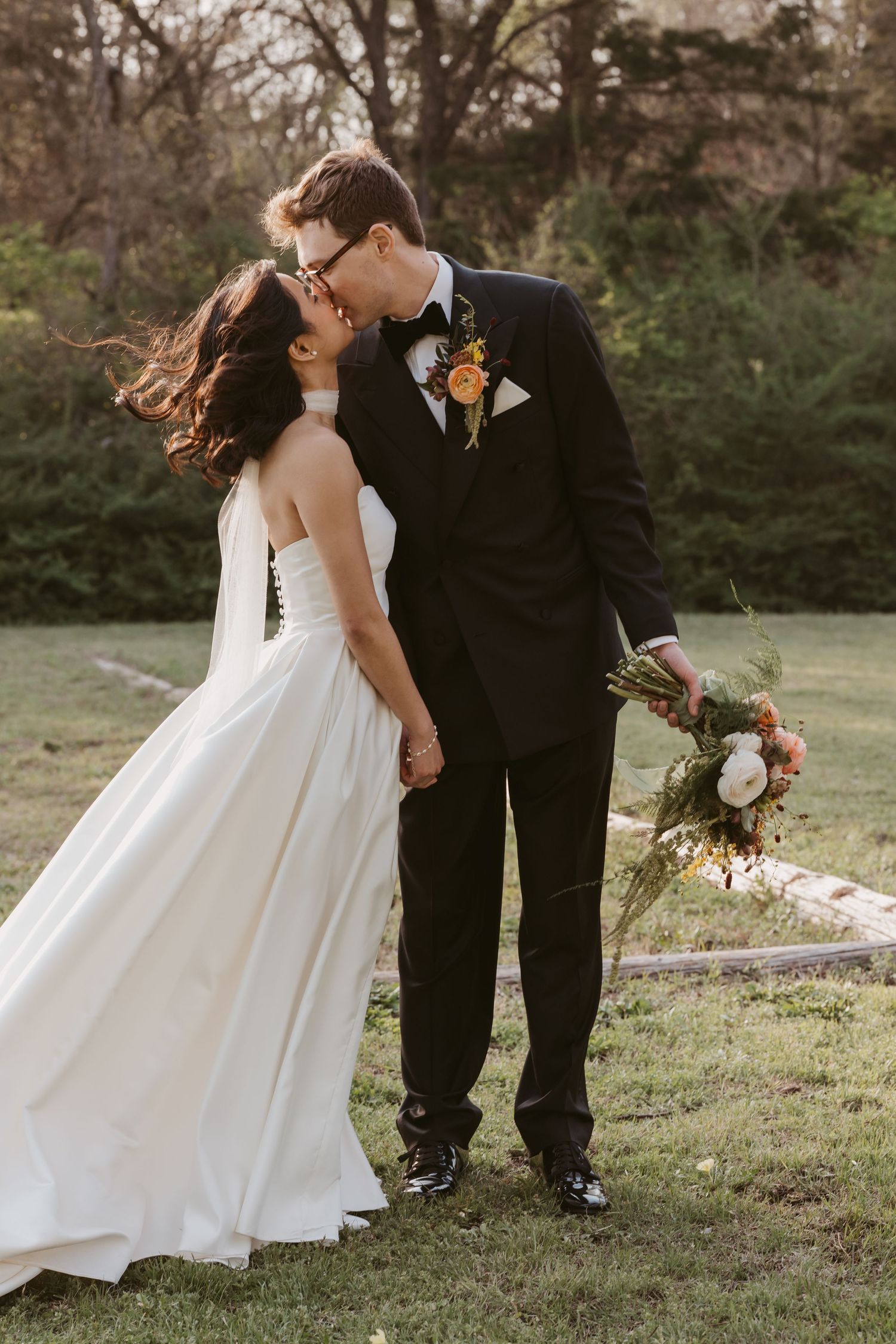 Bride and groom sharing a kiss outdoors, she in a white gown, he in a black tuxedo holding a colorful bouquet.