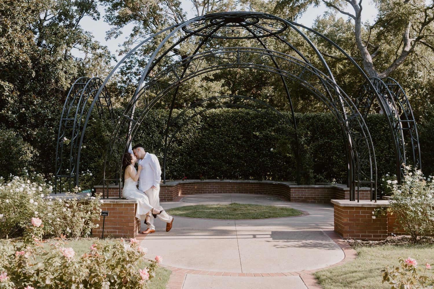 Romantic garden wedding ceremony setup with white floral arch and wooden benches under lush trees.