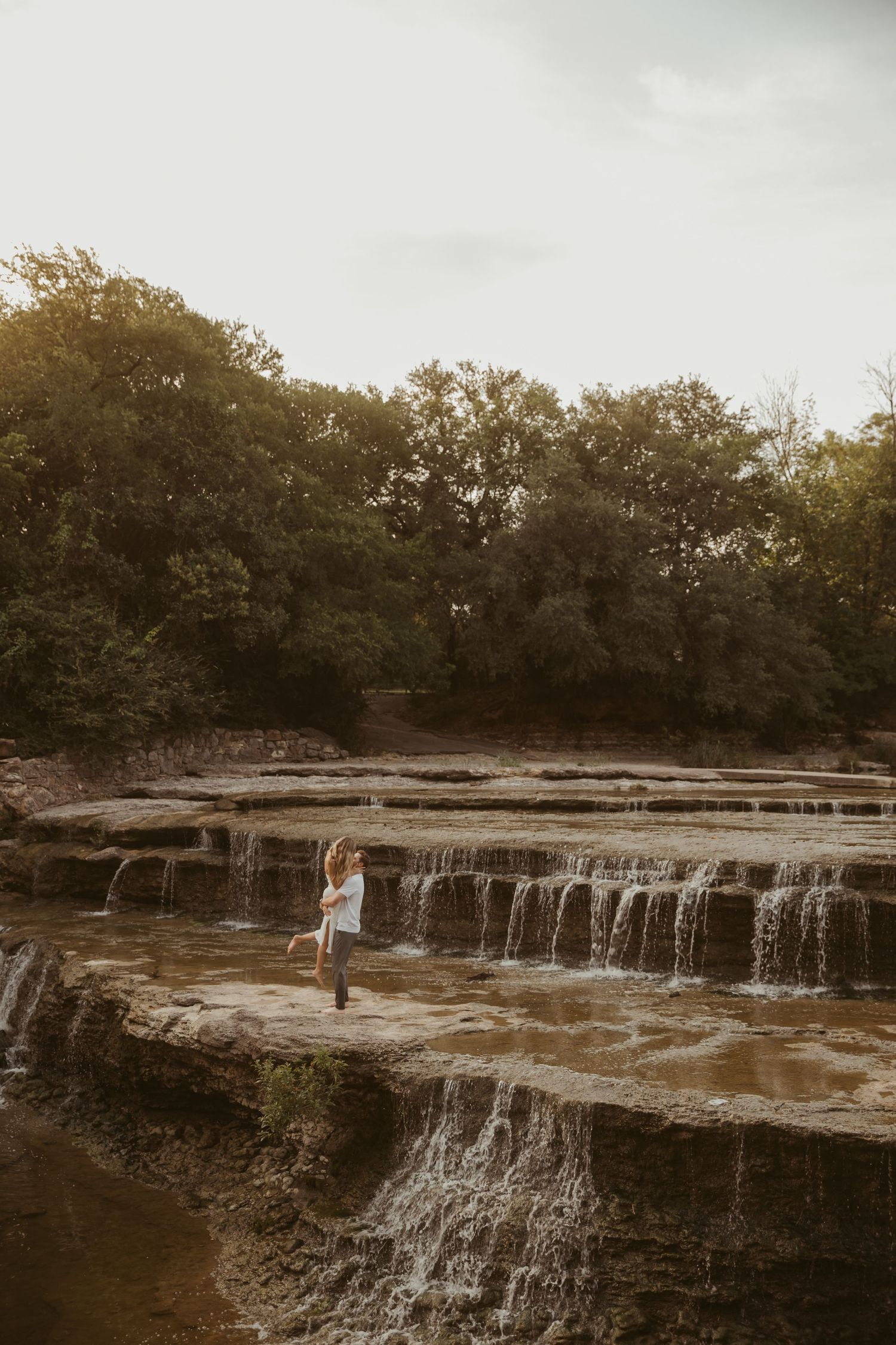 Woman in white dress standing on rocky waterfall steps surrounded by lush green trees at golden hour.