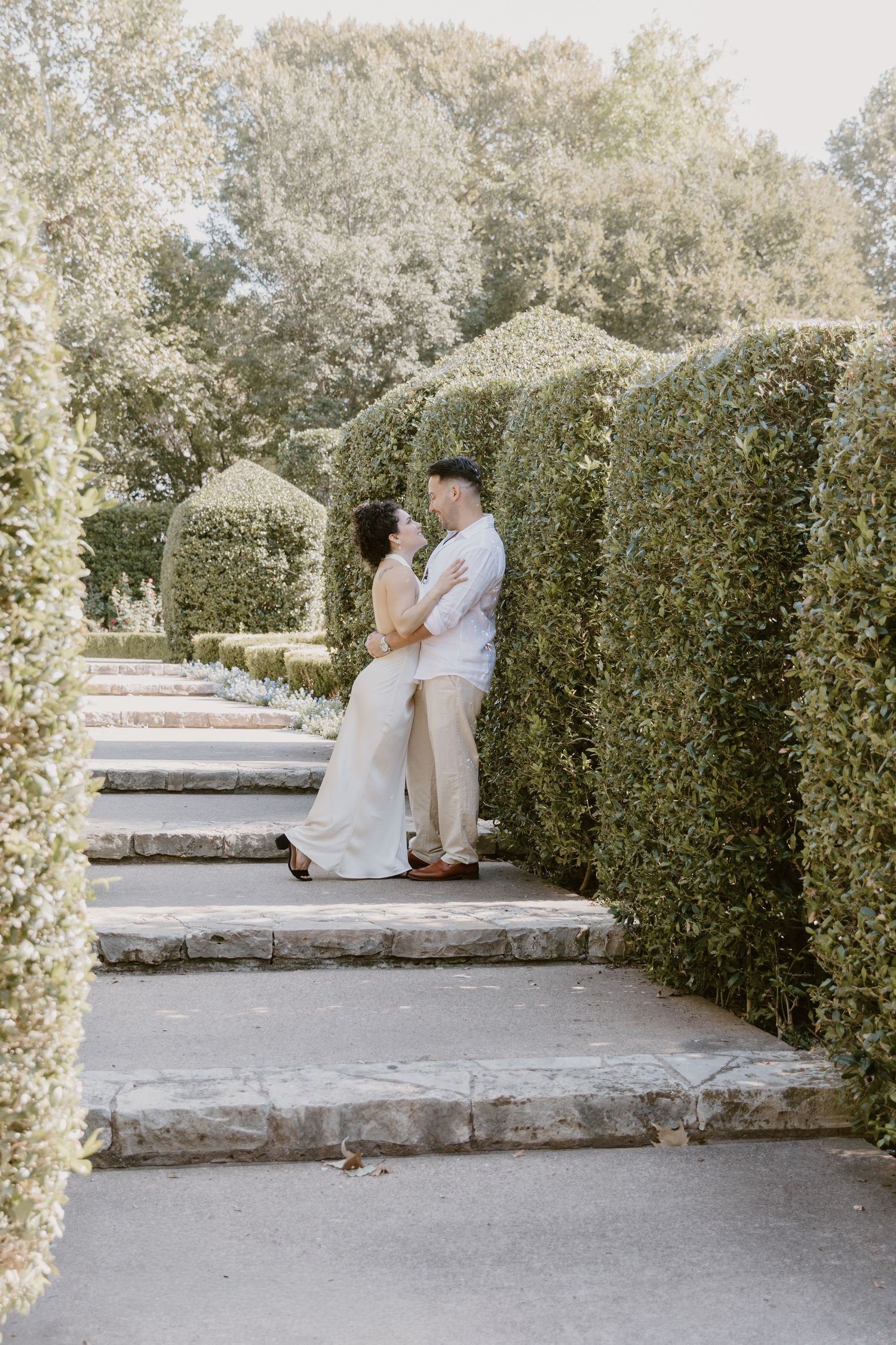 A couple embraces on garden steps surrounded by manicured hedges in an elegant outdoor setting.