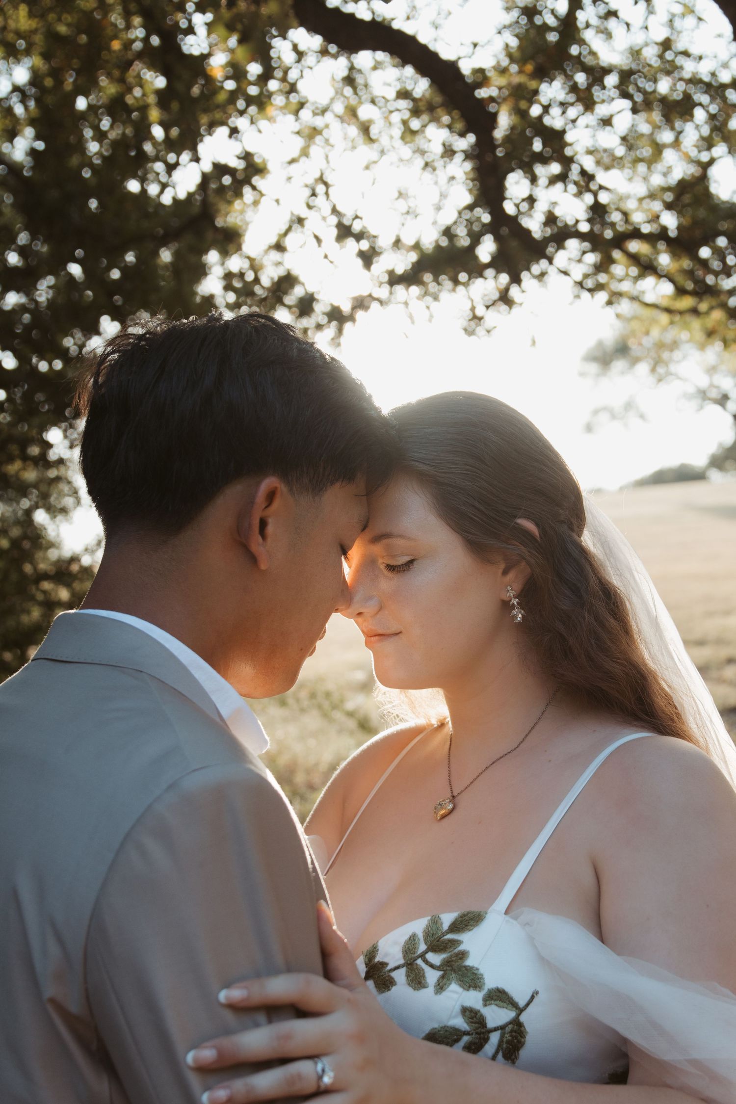 Romantic couple embraces in soft sunlight beneath trees during outdoor wedding photoshoot at golden hour.