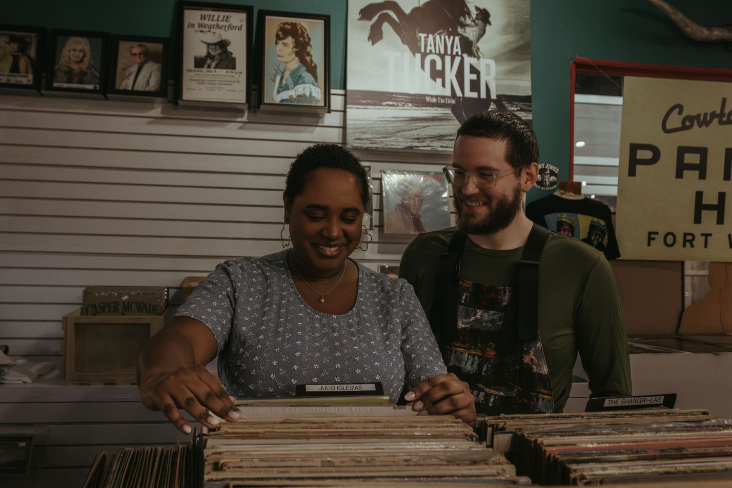 Two people browse through vinyl records together at a retro music store with vintage posters on the wall.