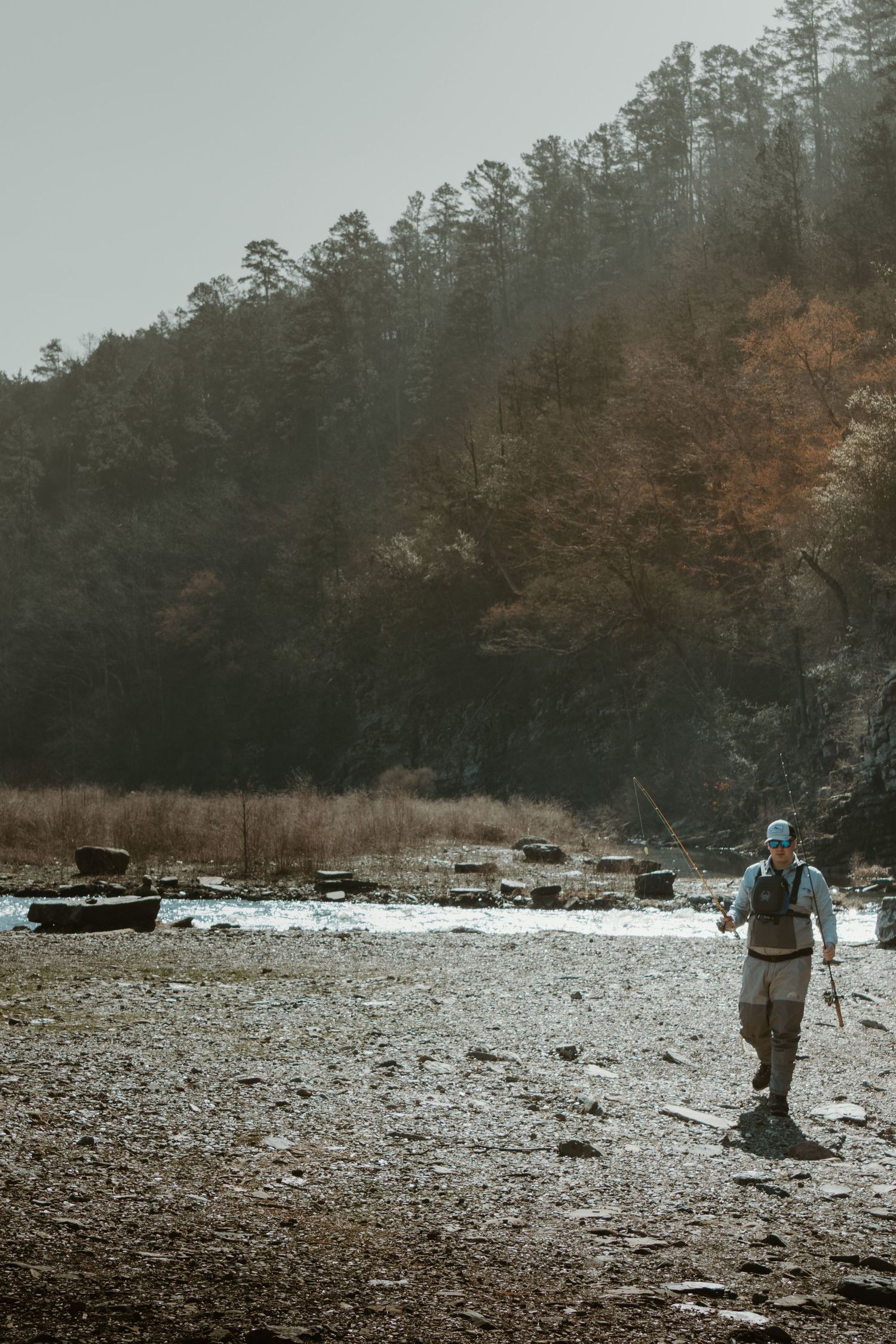 A figure in fishing gear walks along a rocky shoreline beneath a forested hillside.