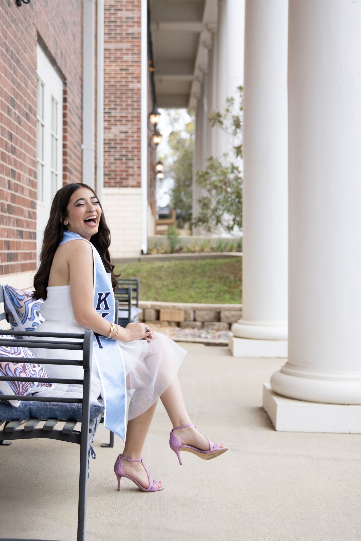 A graduate sits on a bench wearing a light blue dress and pink heels near white columns and brick architecture.