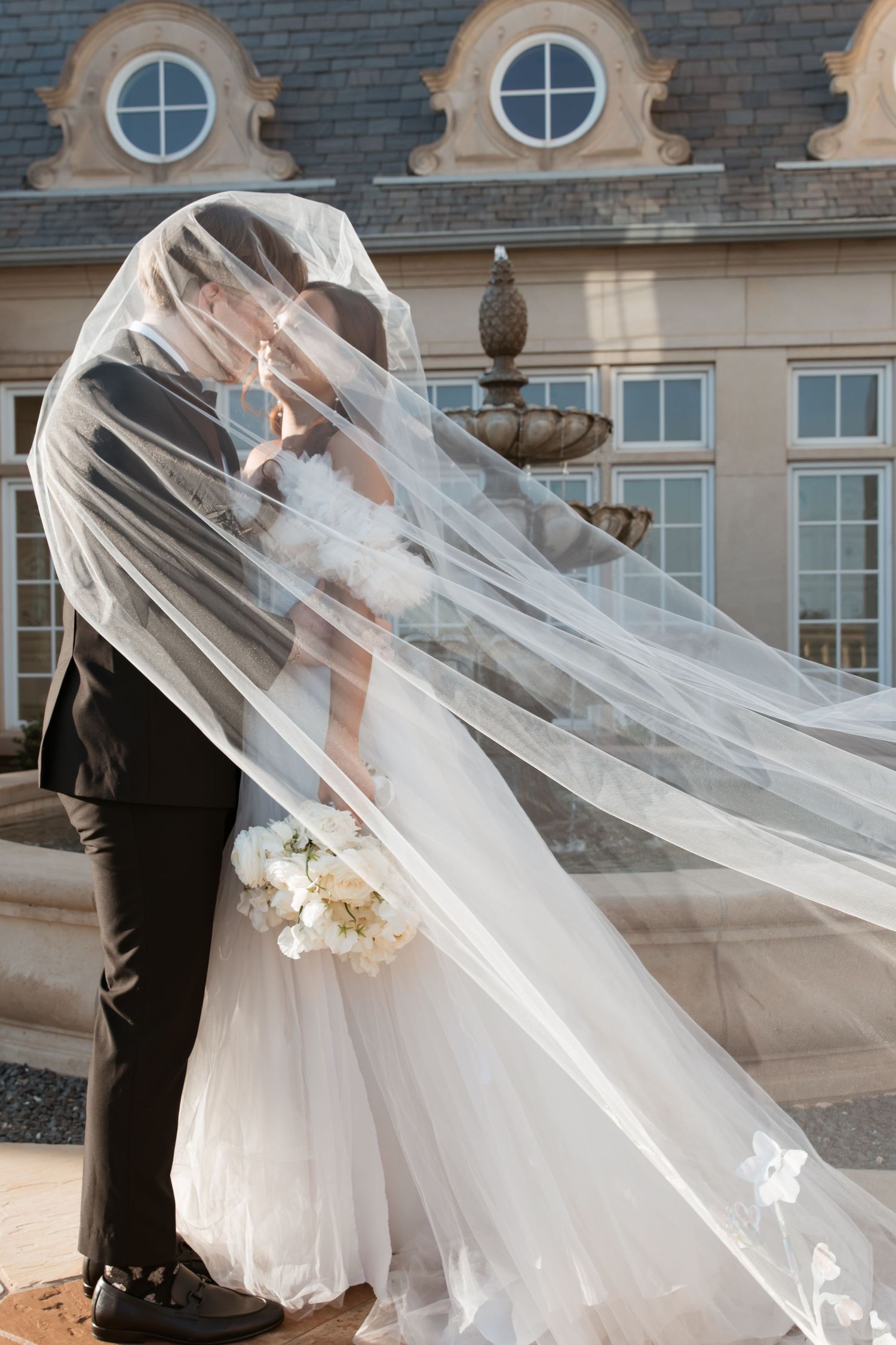 A dramatic cathedral veil billows in the wind as newlyweds embrace in front of a historic building with round windows.