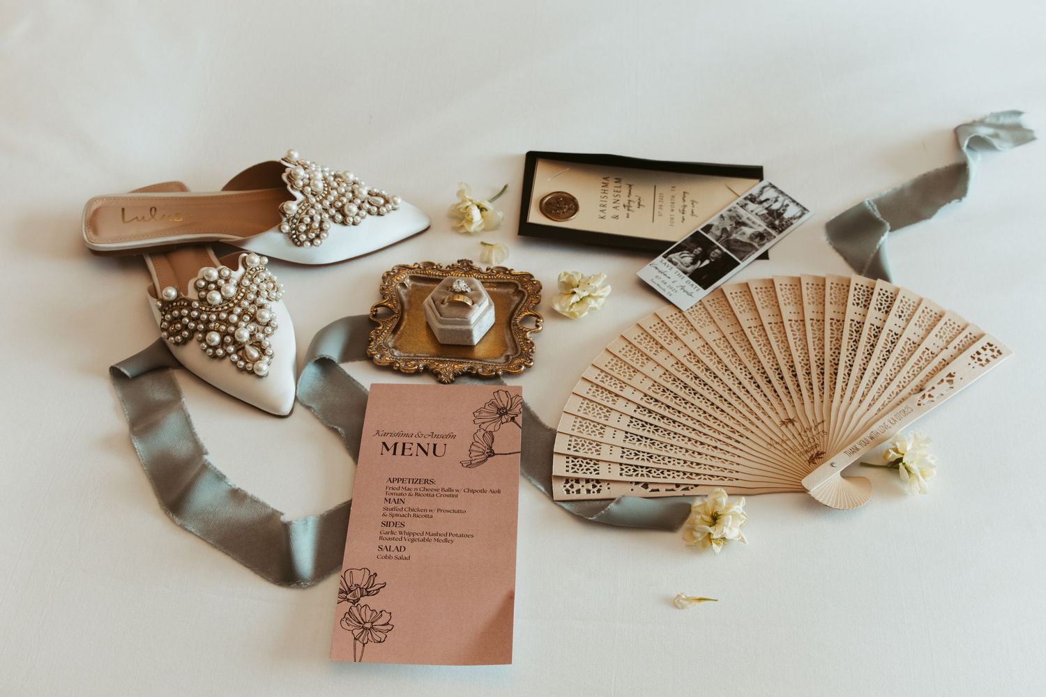Wedding accessories including embellished shoes, a decorative fan, menu cards and ribbons arranged for a photoshoot.