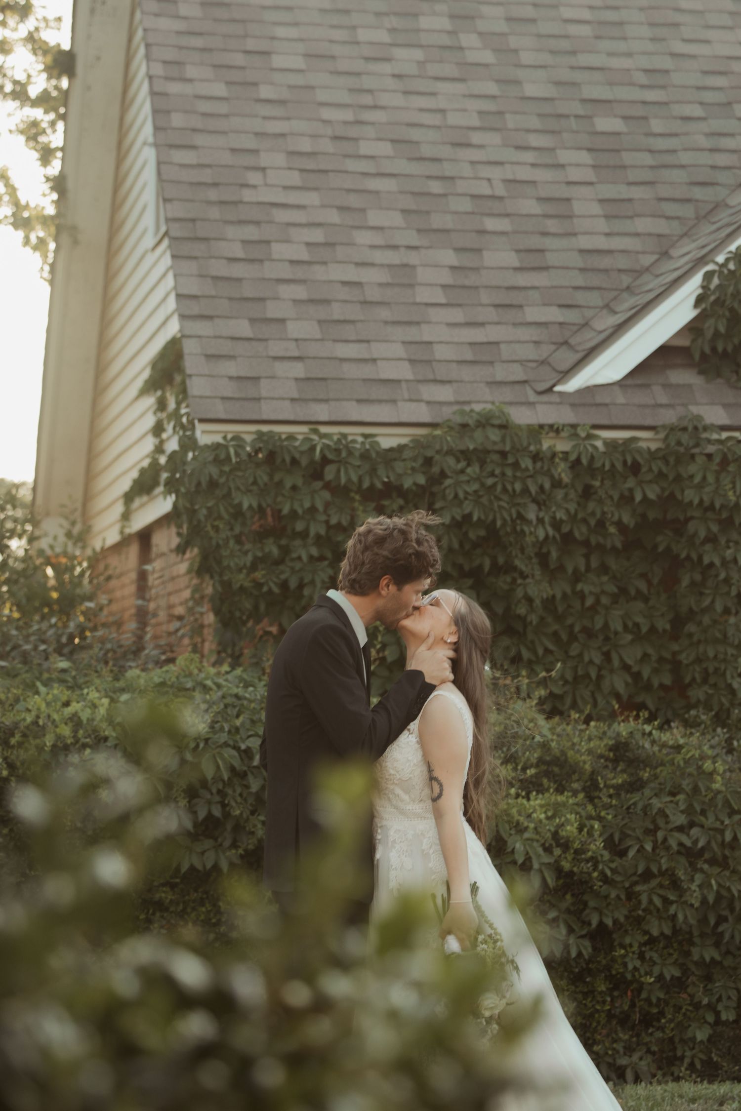 A bride and groom share a romantic kiss in a lush garden setting outside a brick home on their wedding day.